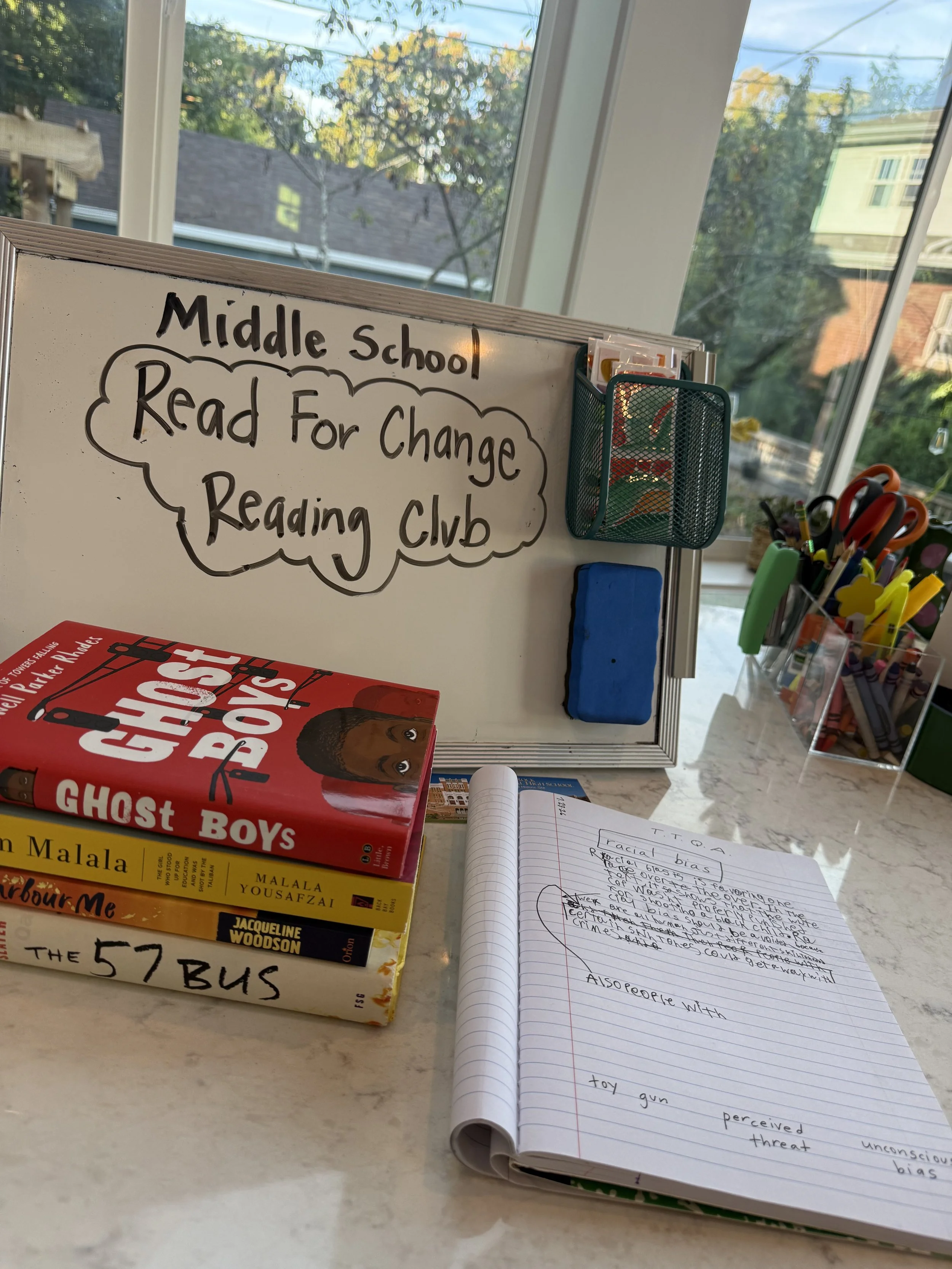 A whiteboard with the words "Middle school Read For Change Reading Club" written on it. Nearby are books, notebooks, and writing utensils, with a window showing trees and rooftops outside.