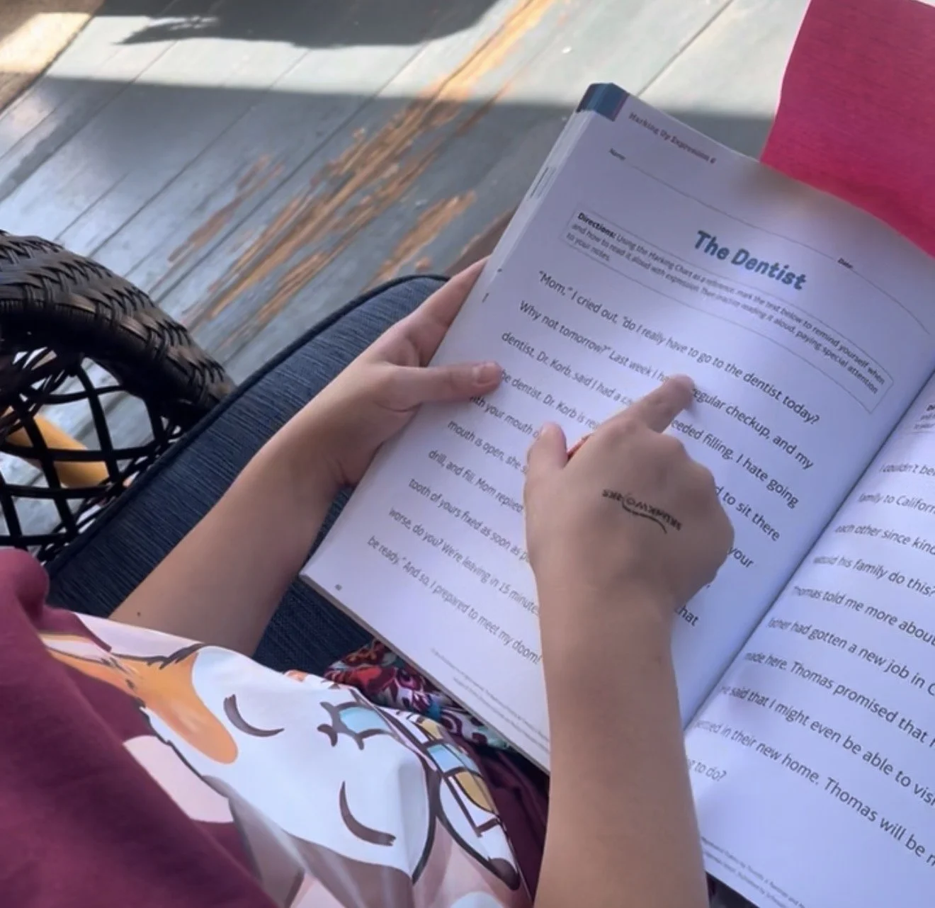 Person sitting outdoors on a bench, reading a book, with a wooden deck floor visible. The person is wearing a pink top and floral patterned pants.