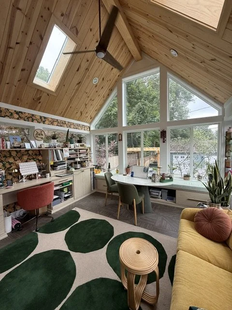 A cozy attic workspace with large windows, a yellow sofa with pink pillow, a white desk with three chairs, a green and white patterned rug, a built-in bookshelf, and various decorative plants and items.