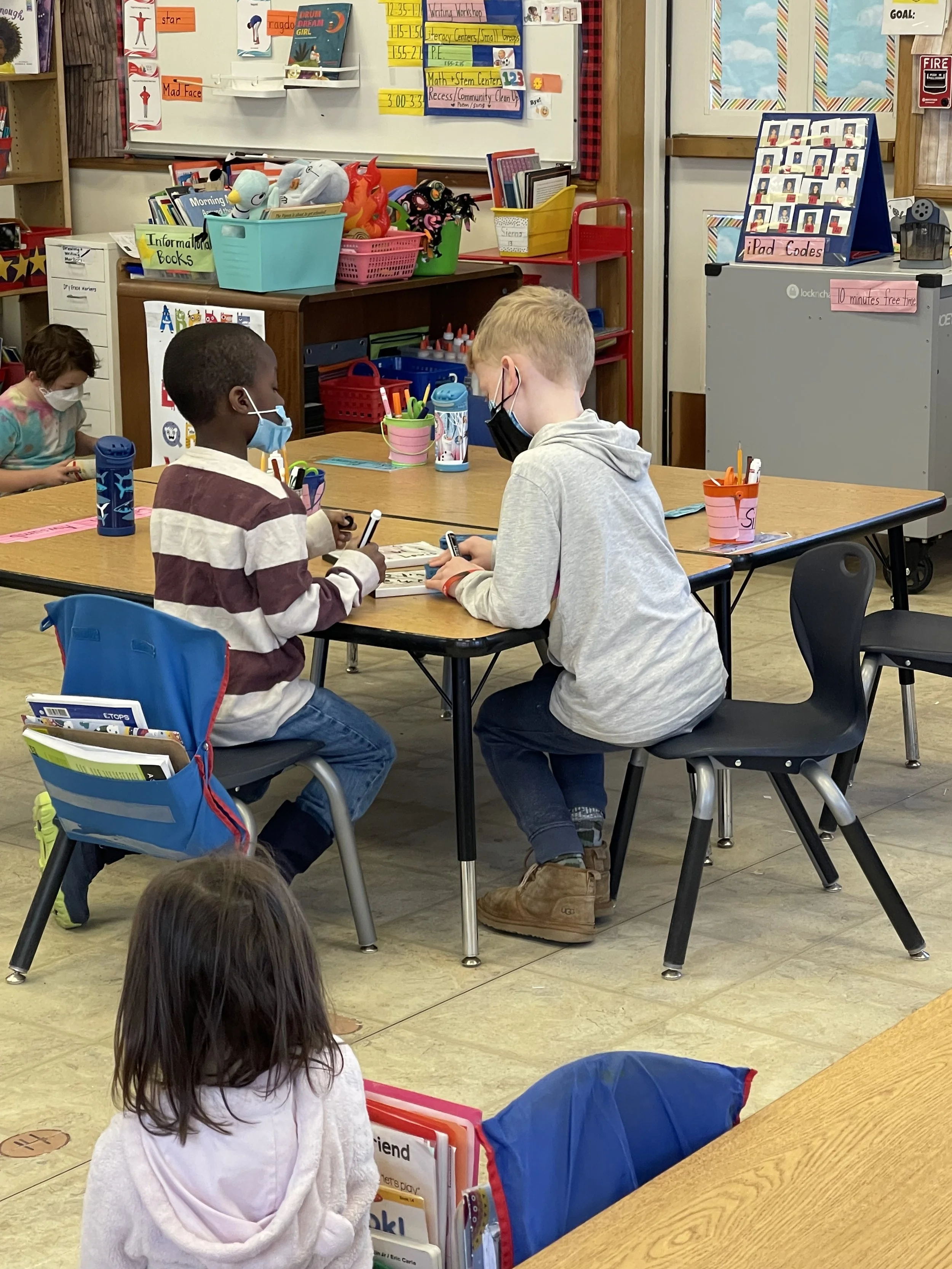 Two young boys wearing face masks are talking at a table in a classroom, with a girl sitting nearby. The classroom has educational materials, colorful bins, and posters on the walls.