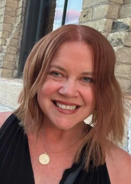 A woman with shoulder-length red hair, smiling, wearing a black top and a gold pendant necklace, standing outside near a brick wall and window.