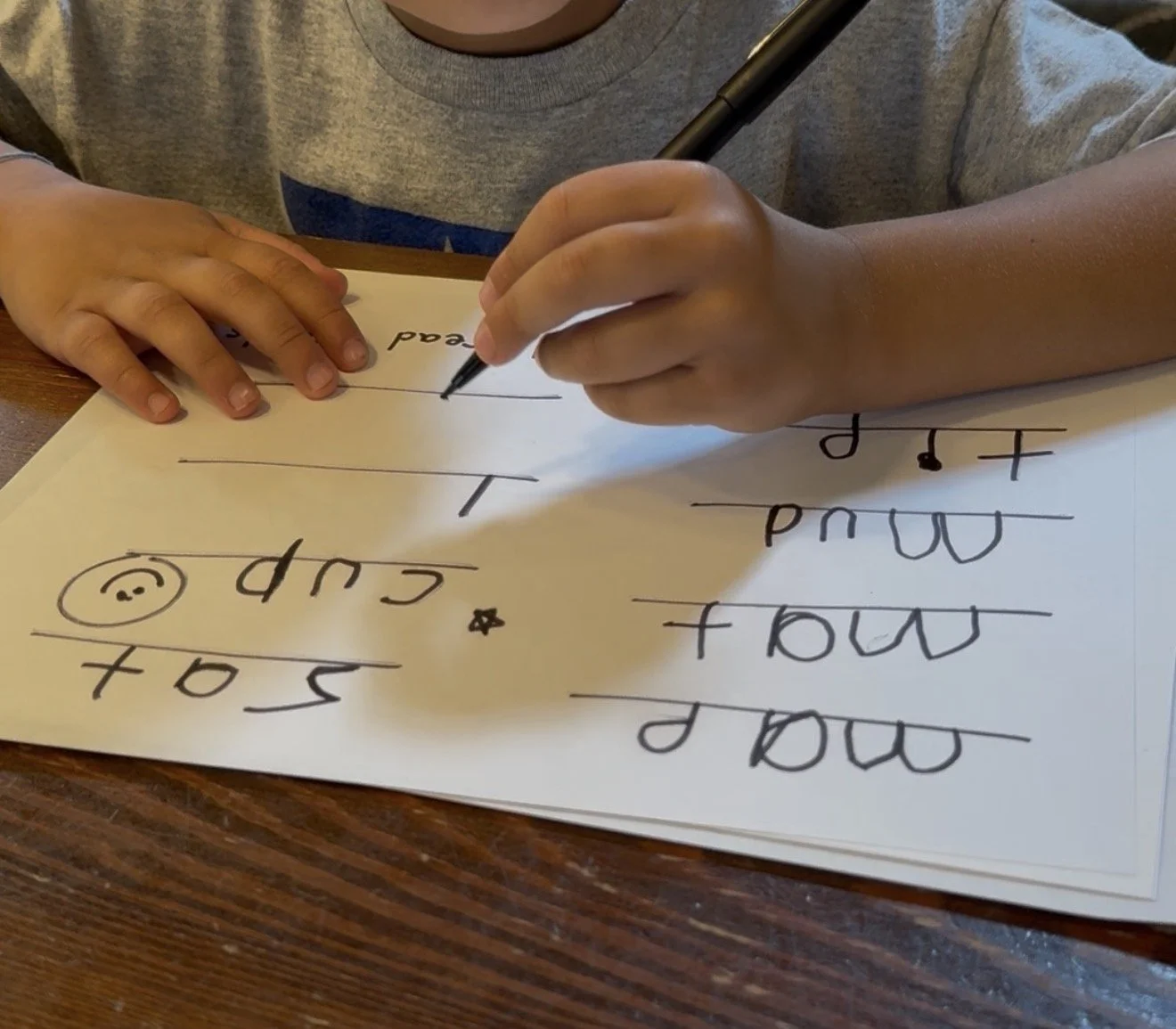 A child on a wooden table practicing cursive writing on a large sheet of paper, with their hand holding a black marker.