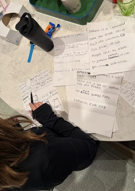 A person working on handwritten notes and sketches about Jamaica at a table with papers, a black water bottle, a blue pencil, and a green container. The person is writing with a black pen.