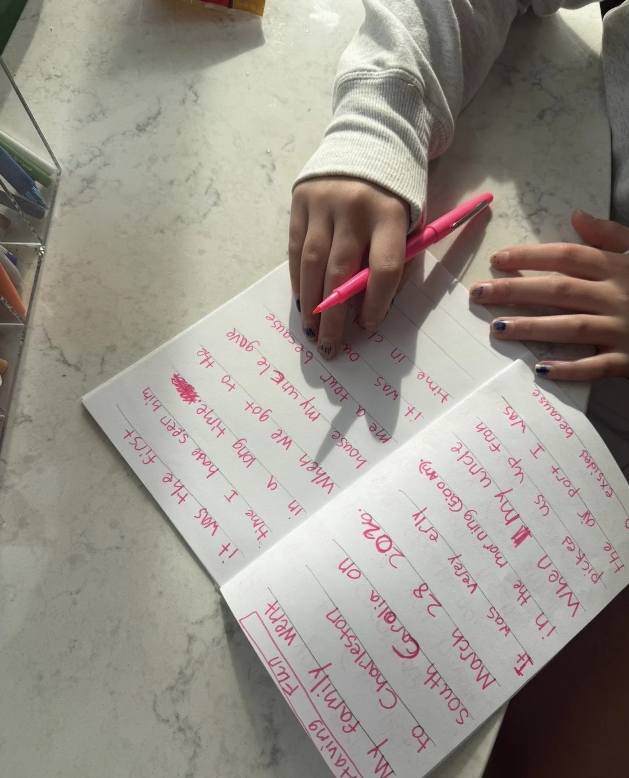 A person writing in a notebook with a pink pen. The notebook contains handwritten notes in pink ink. The person is wearing a beige sweatshirt and has painted fingernails. The surface is a light-colored marble countertop with a plastic organizer on the side.