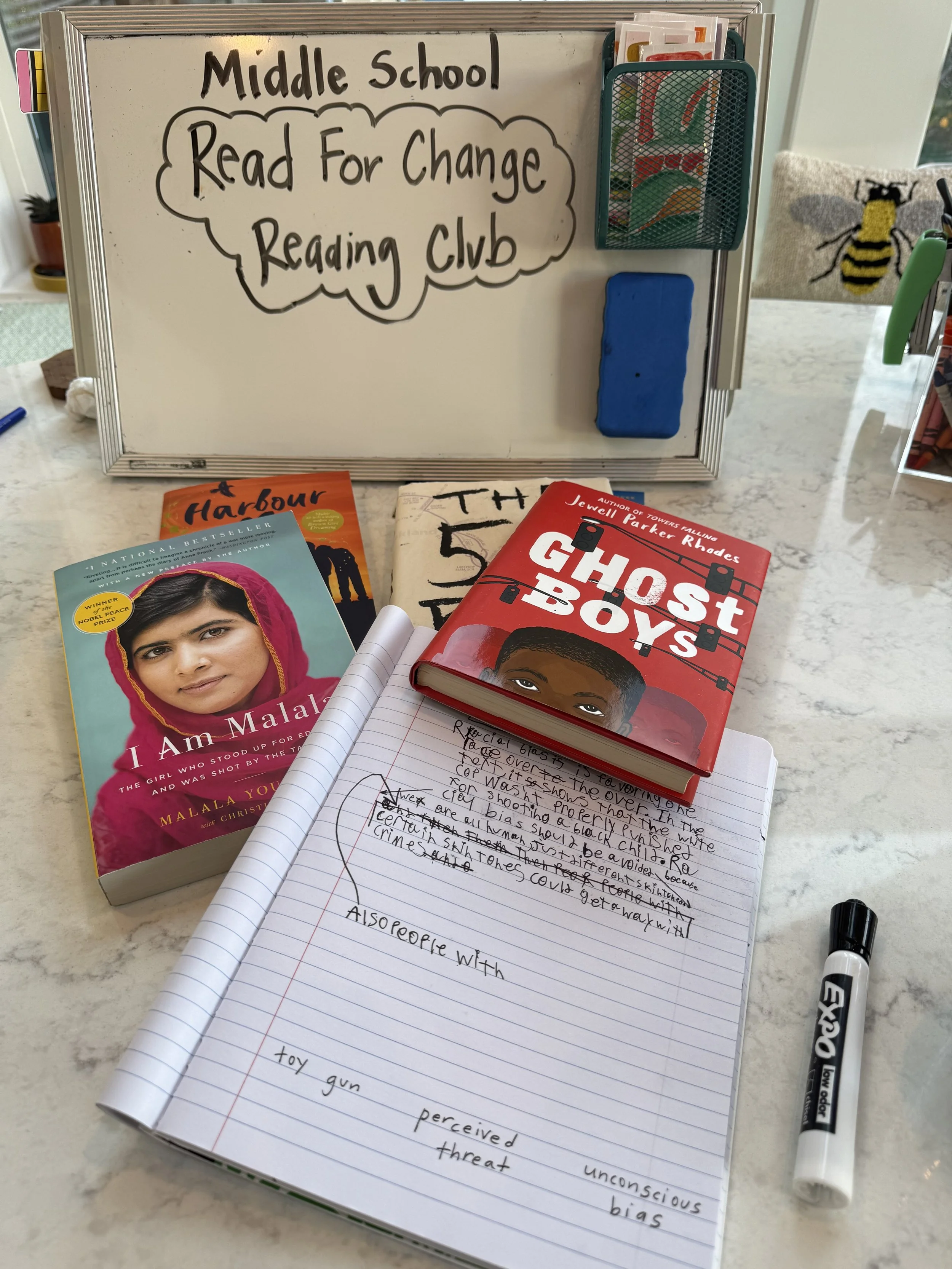 Whiteboard with the words 'Middle School Read For Change Reading Club' written on it, a pen holder with markers, and books including 'I Am Malala' and 'Ghost Boys' placed on a marble table. An open notebook with handwritten notes is also visible.