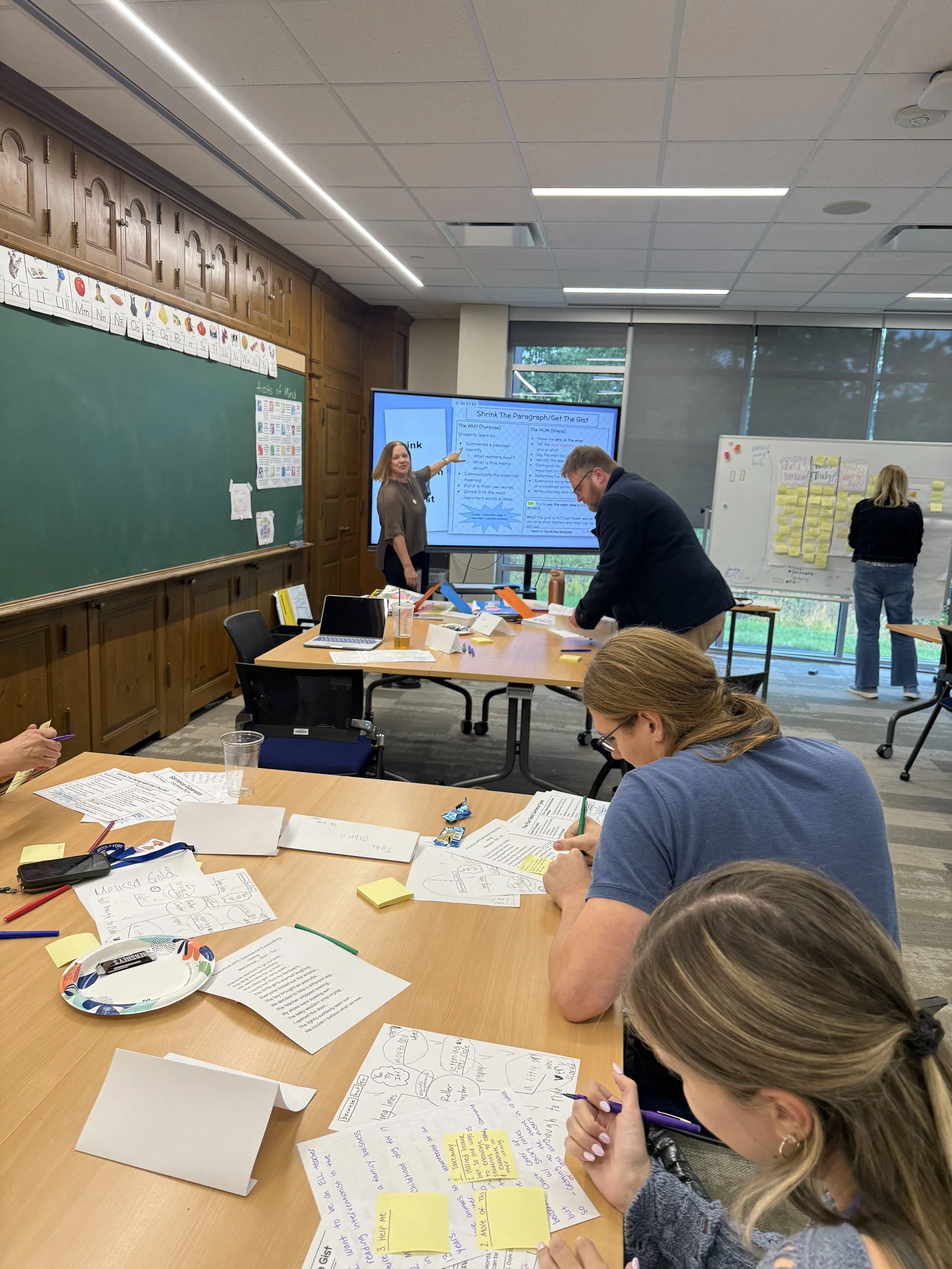 A group of people in a classroom engaged in a workshop or collaborative activity. Some are writing or drawing on papers with notes and diagrams. A woman is pointing at a large screen displaying a presentation. The room has a green chalkboard, a white
