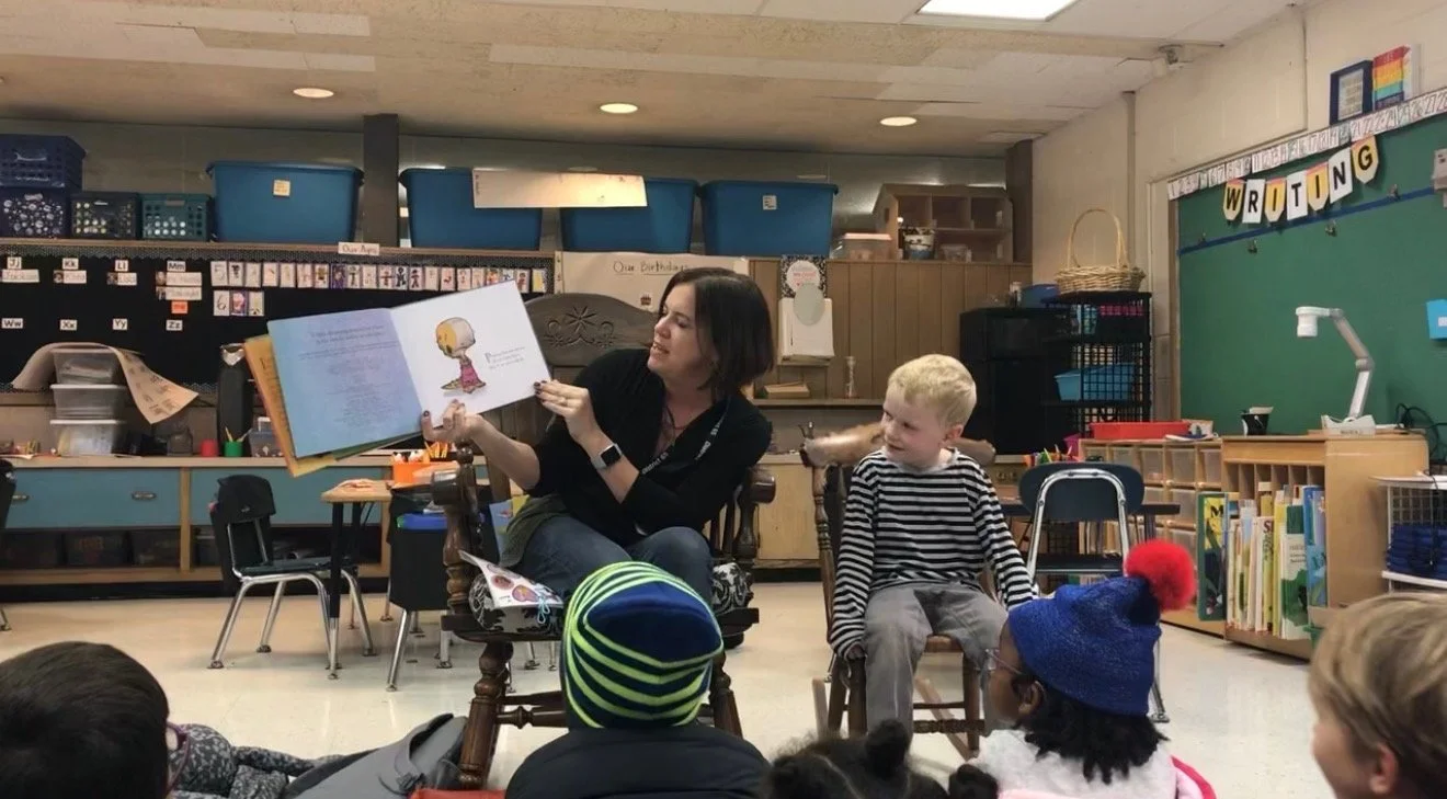 A woman reading a picture book to a group of children in a classroom. The woman is sitting on a rocking chair, and the children are sitting on the floor and small chairs, attentively listening.