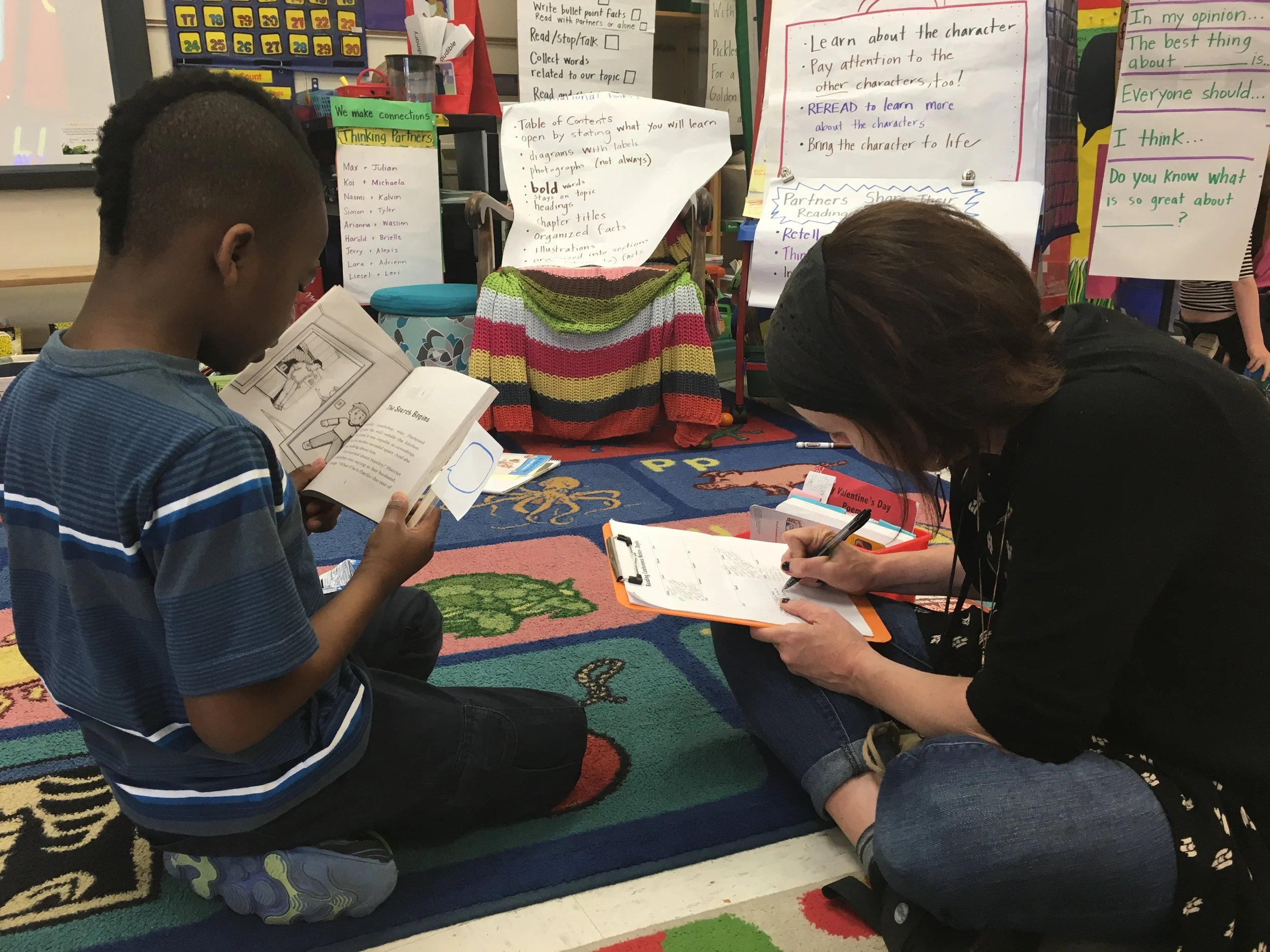 A classroom setting with two children sitting on the colorful carpet, reading and taking notes. The background contains various posters, charts, and teaching materials, including a chart labeled 'Table of Contents' and another poster with writing pro