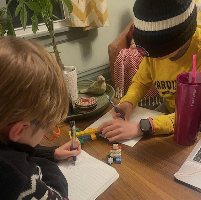 Two children sitting at a table, writing on notebooks with pens, surrounded by school supplies and a small decorative bird statue.