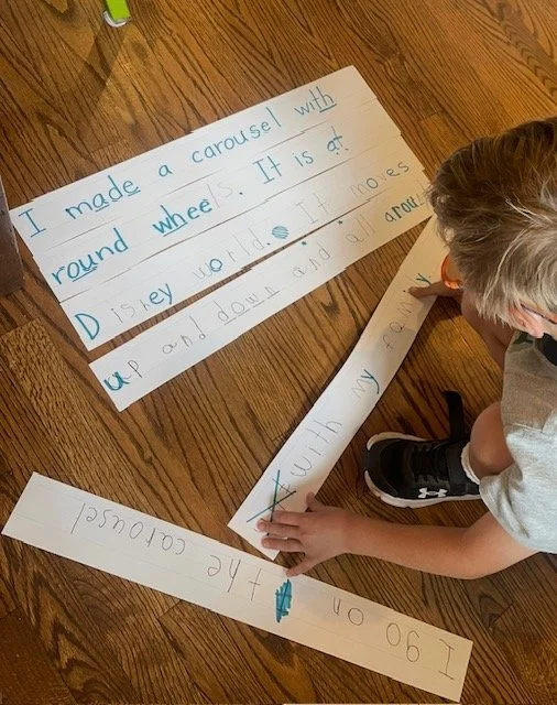 A child with gray hair wearing a gray t-shirt and black sneakers is working on a school project on a wooden floor. There are several long strips of white paper with handwritten text in blue ink, describing making a carousel with a round wheel and Dis
