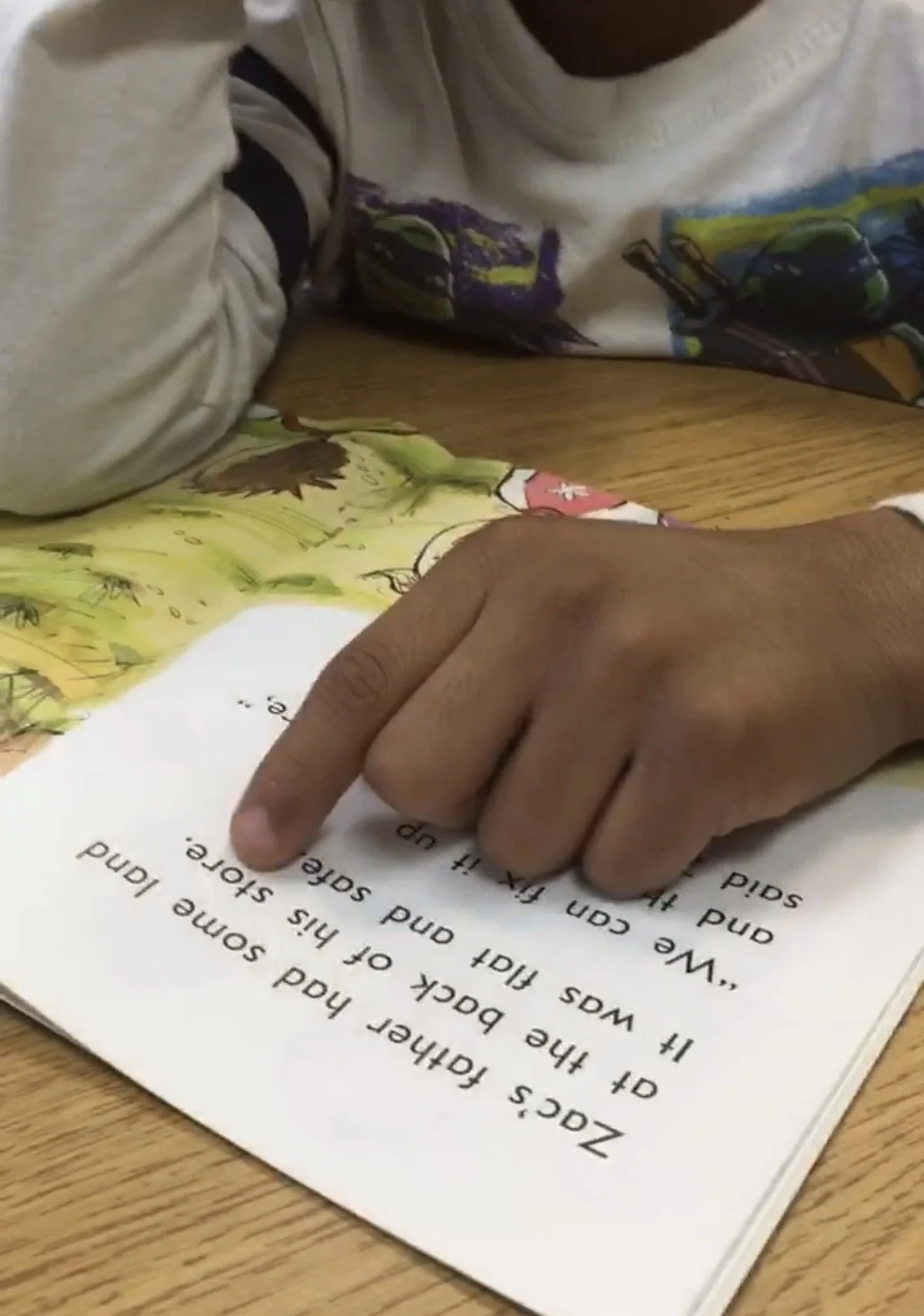 A person reading a book with a colorful illustration of vehicles on the page, pointing at the words with their finger, on a wooden table.