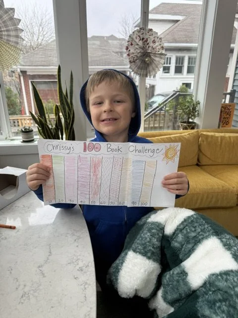 A young boy smiling and holding a colorful chart titled 'Chrissy's 100 Book Challenge' inside a bright room with large windows, plants, and a yellow couch.