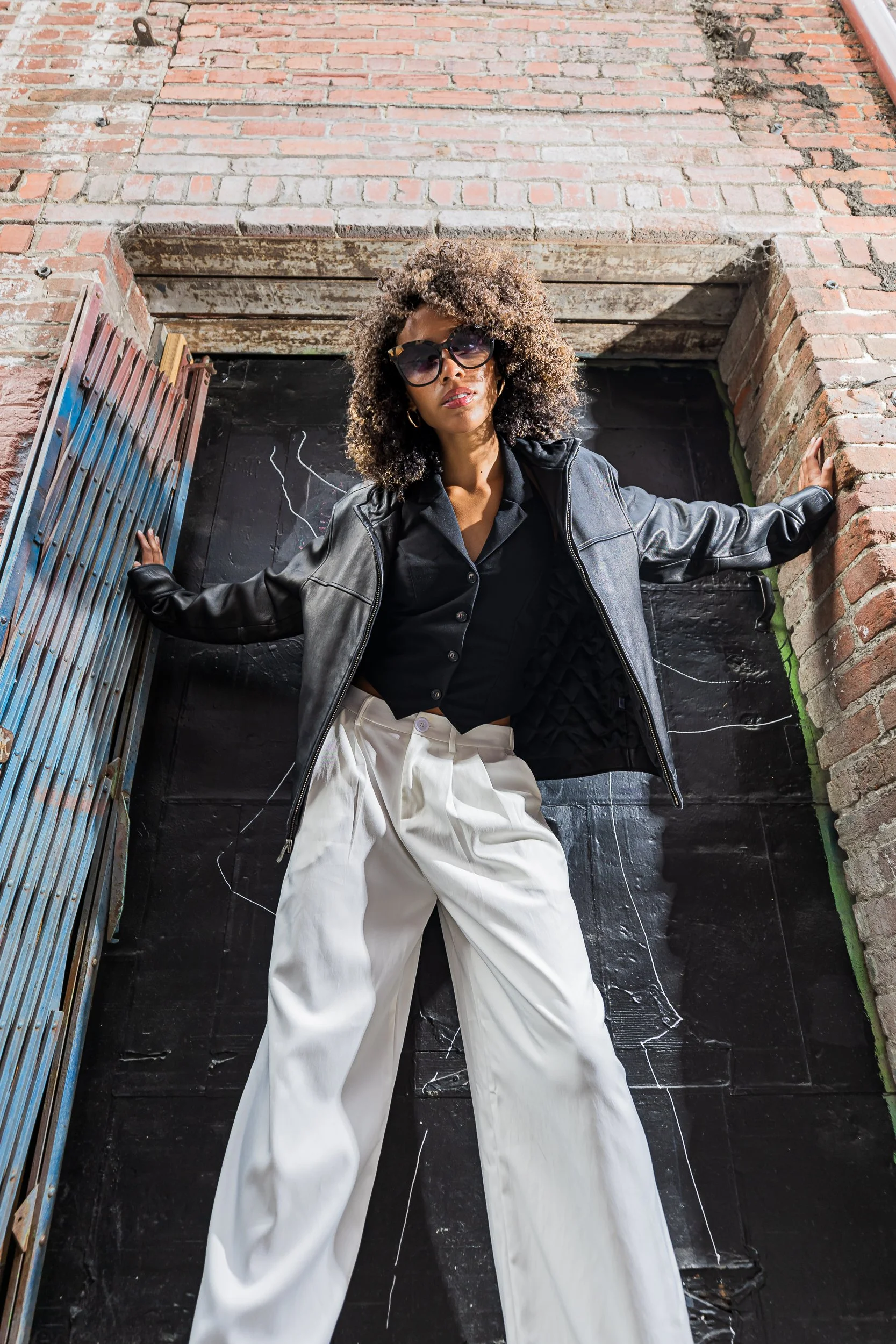 Model in a leather jacket and white wide leg pants posing in a brick doorway with a dark metal door