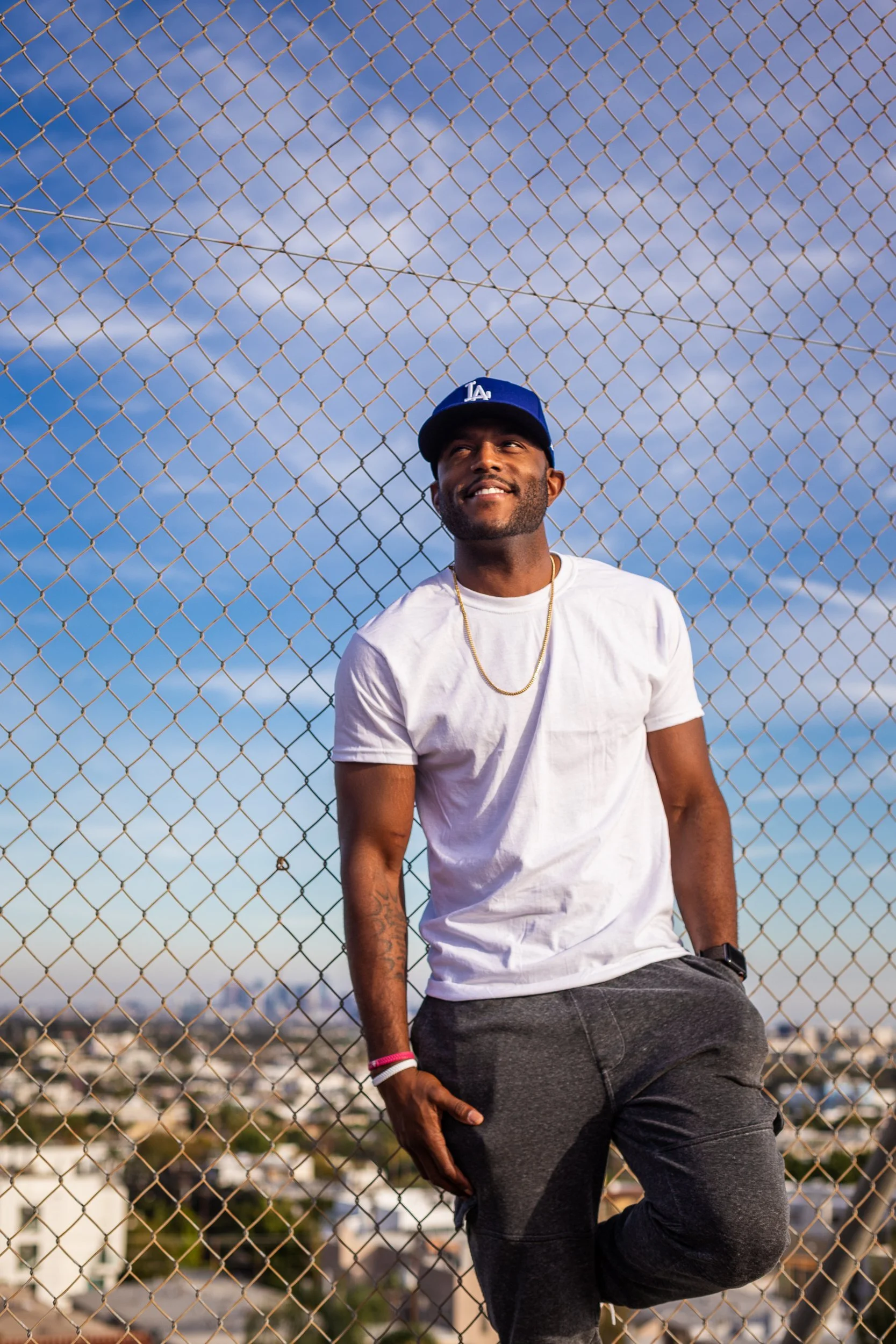 Man in a white tee, gold chain and blue cap standing in front of a chain link fence under a cloudy blue sky