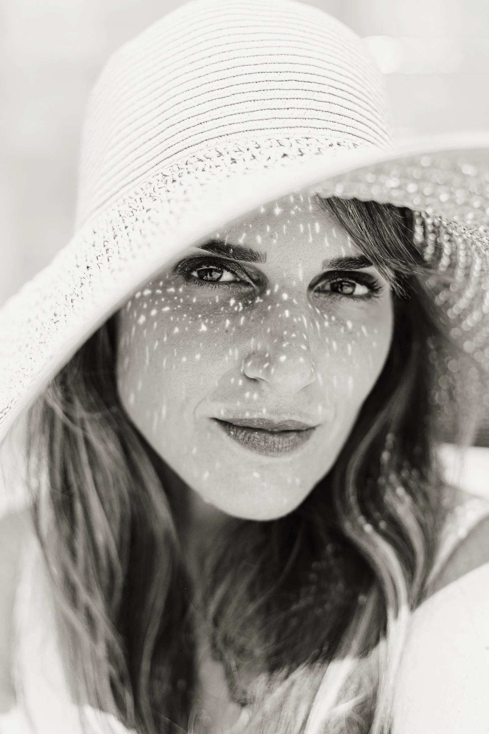 Black and white close portrait of a woman in a wide brim straw hat with dappled light across her face