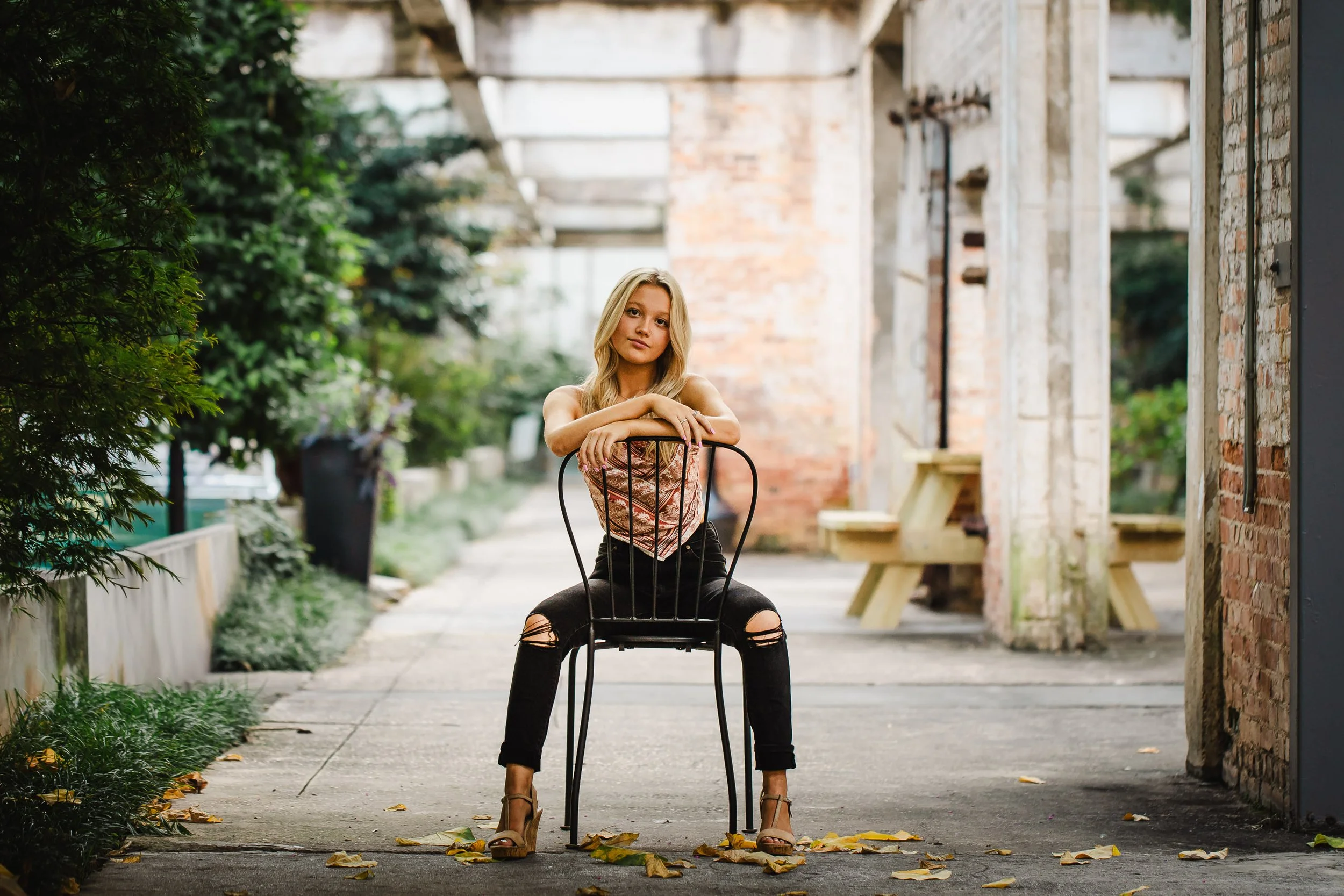 Woman in a white top and ripped black jeans sitting on a black metal chair in a brick walkway with fallen leaves