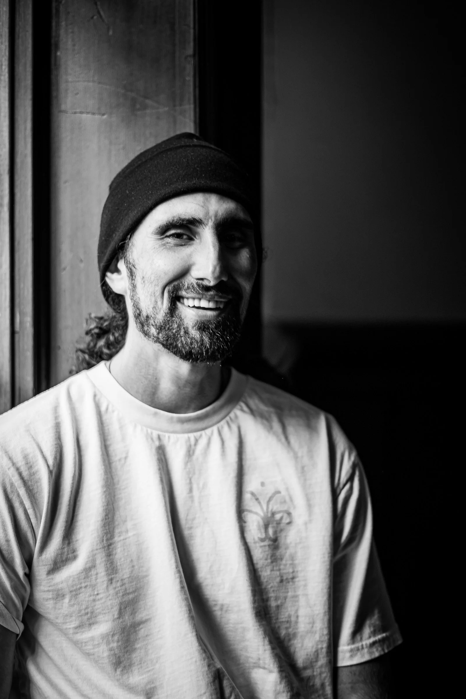 Black and white portrait of a smiling man in a beanie and white tee seated by a window