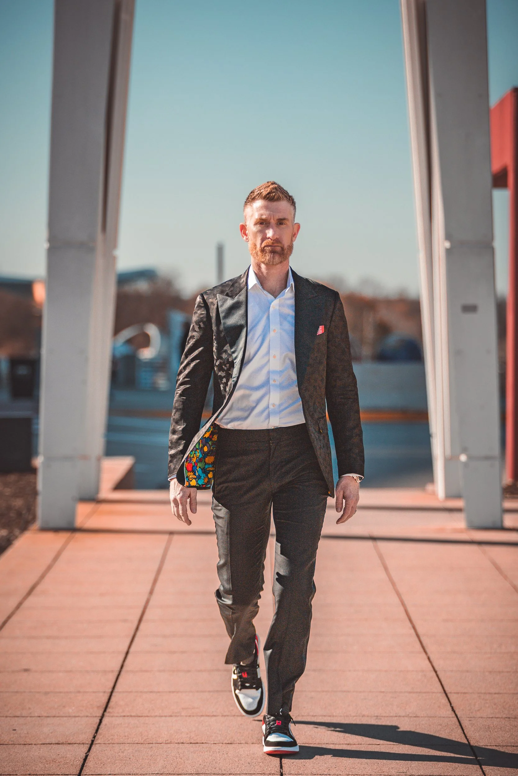 Man in a charcoal suit and white open collar shirt walking along a peach colored walkway between concrete columns at golden hour