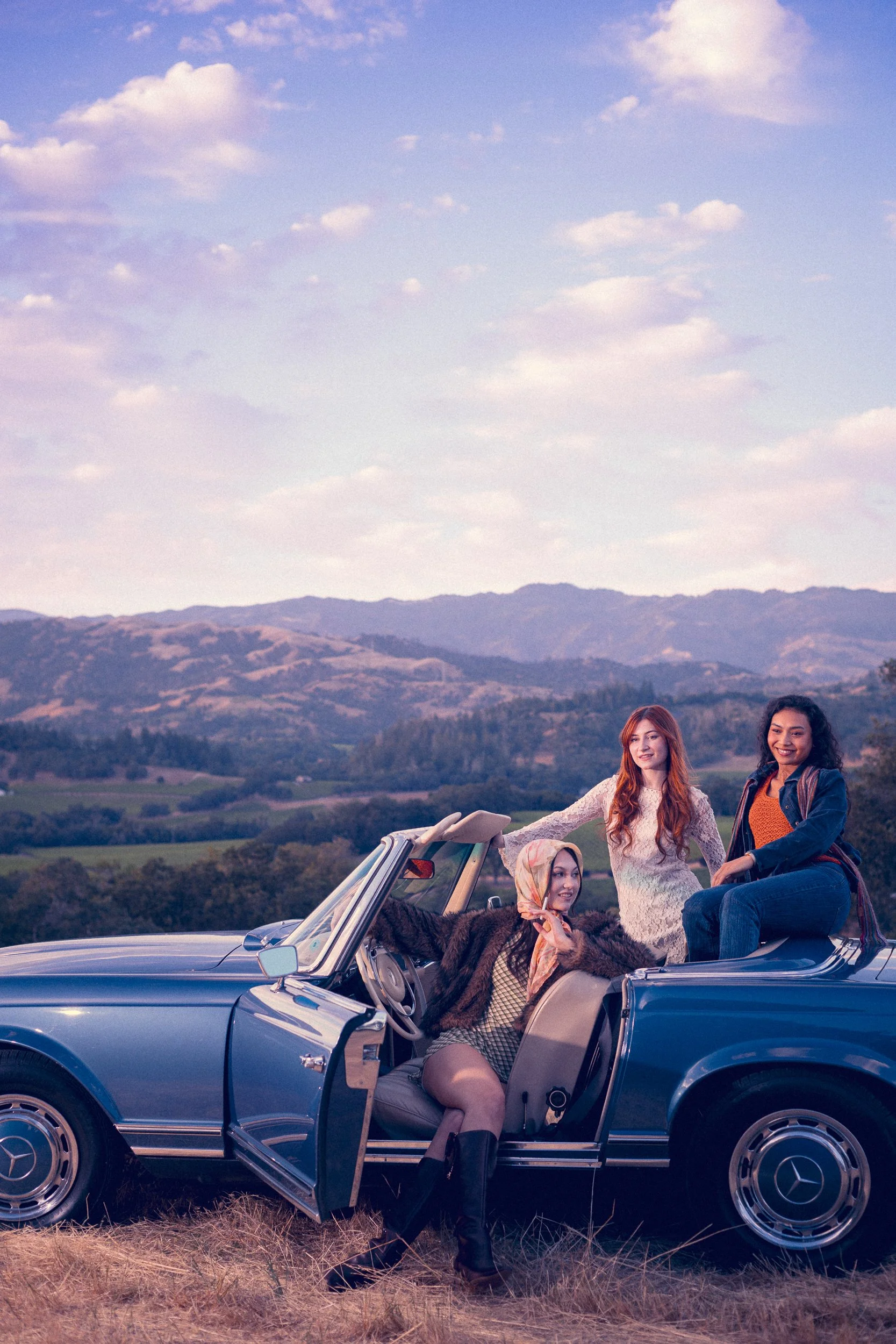 Three women posing on and beside a vintage blue Mercedes convertible at dusk with hills and a pastel sky behind them