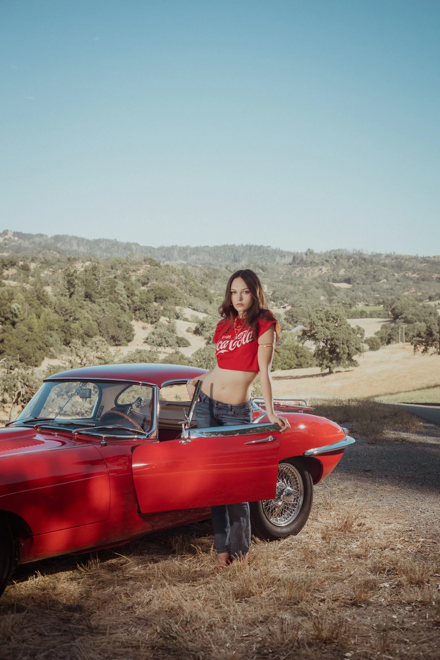 Woman in a red cropped tee standing beside the open door of a vintage red sports car on a country road with golden hills behind