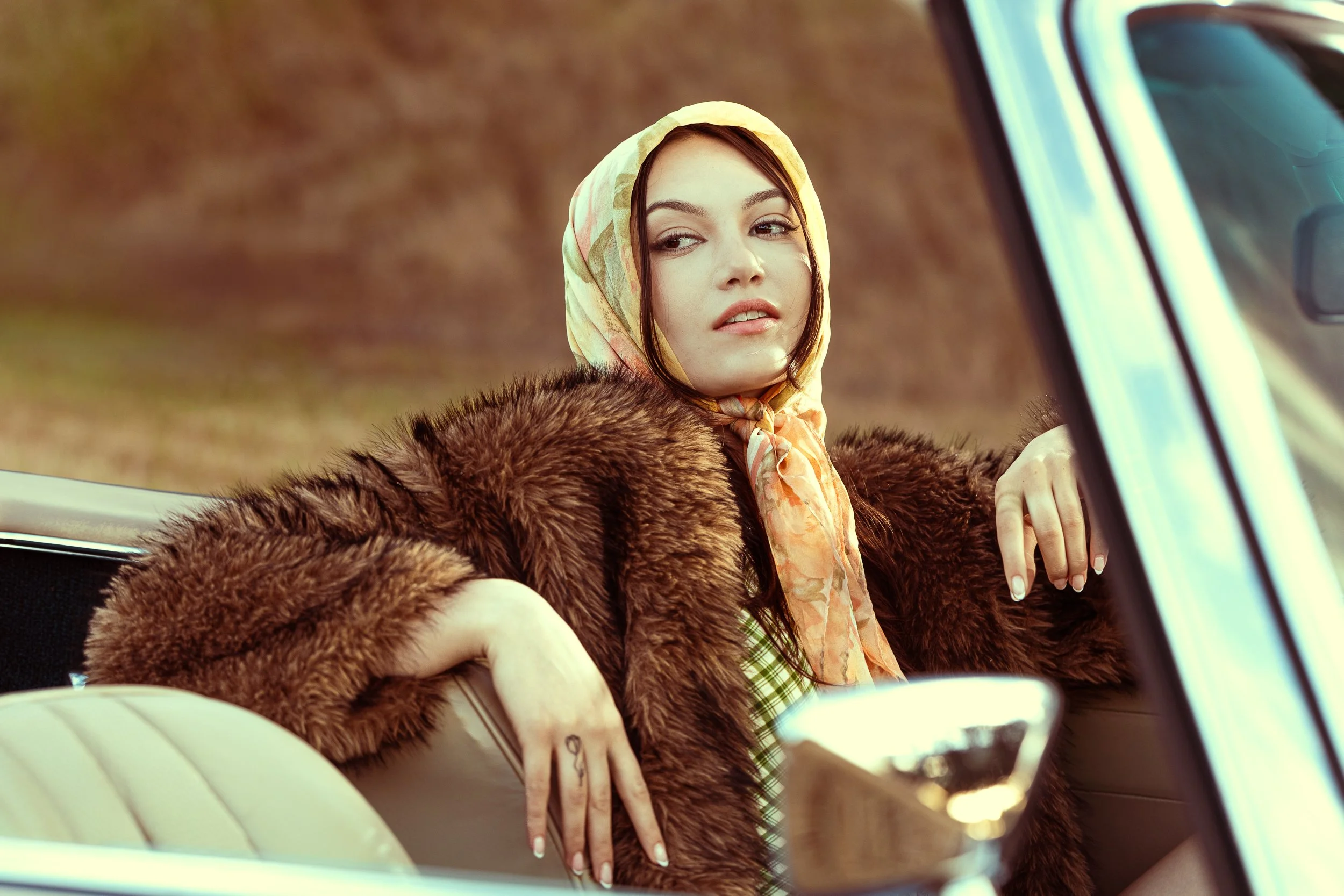 Woman in a silk headscarf and fur coat resting her arm on the seat of a vintage convertible in warm light