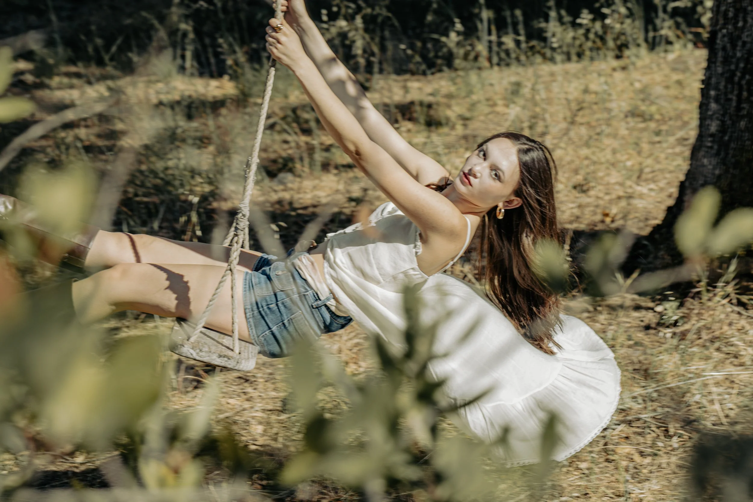 Model in a white tunic, denim shorts and brown boots swinging on a rope swing, looking at the camera through soft green leaves