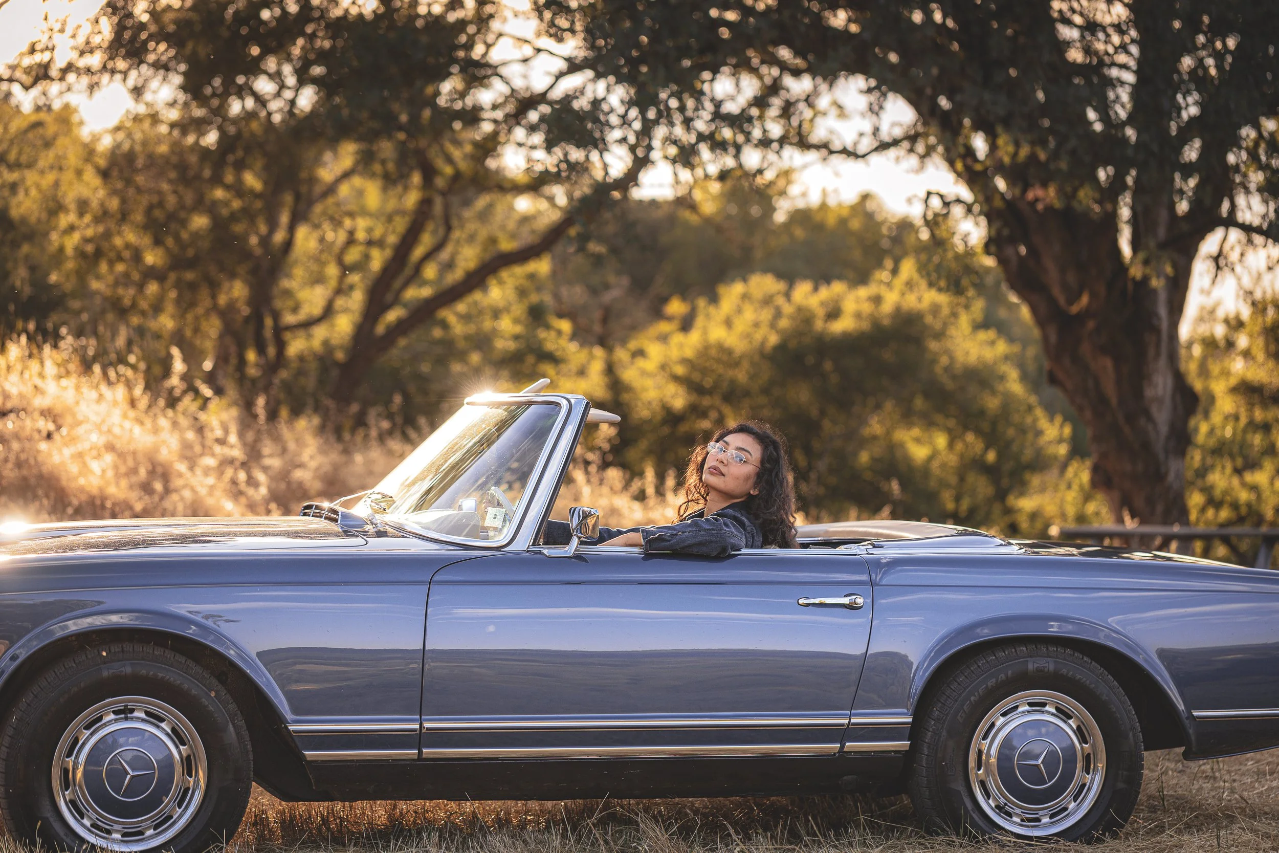 Woman relaxing in a vintage blue Mercedes convertible parked under oak trees in warm golden light