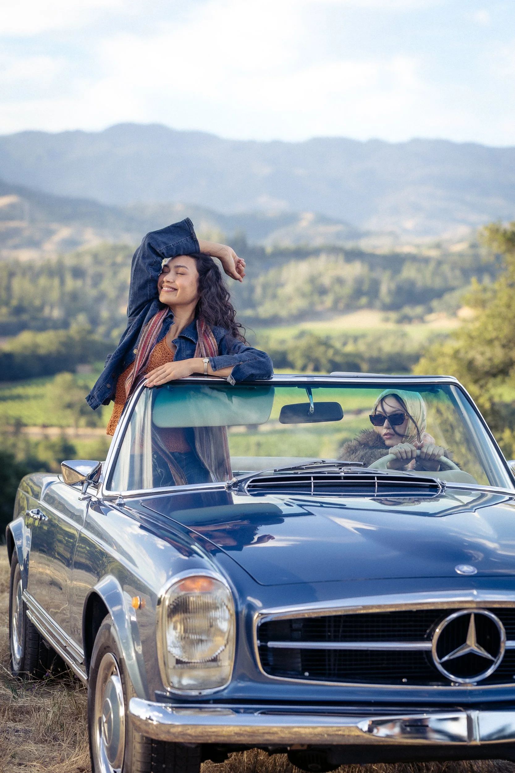 Woman in a denim jacket leaning over the windshield of a vintage blue Mercedes convertible with rolling hills behind her