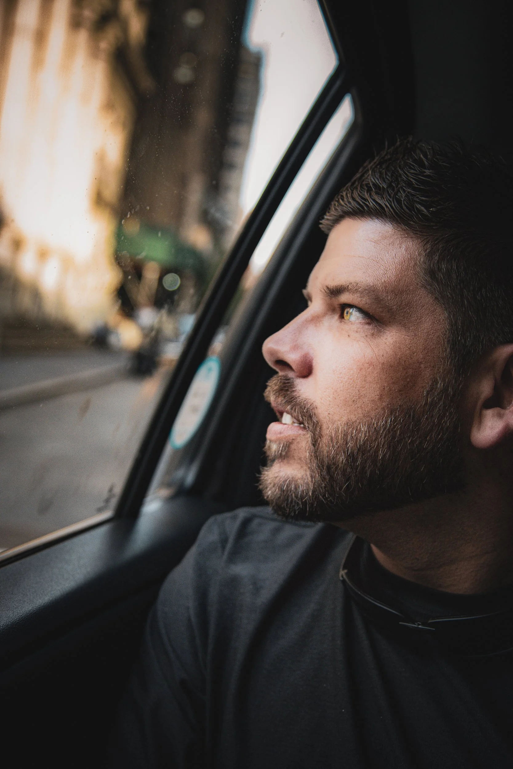 Bearded man in a dark tee looking out of a car window with warm light on his face