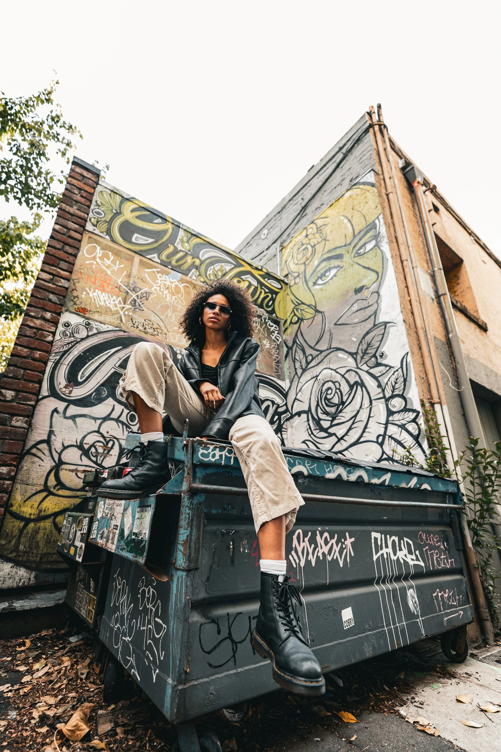 Model in a leather jacket and cream pants sitting on a graffiti covered dumpster, shot from a low angle against a mural