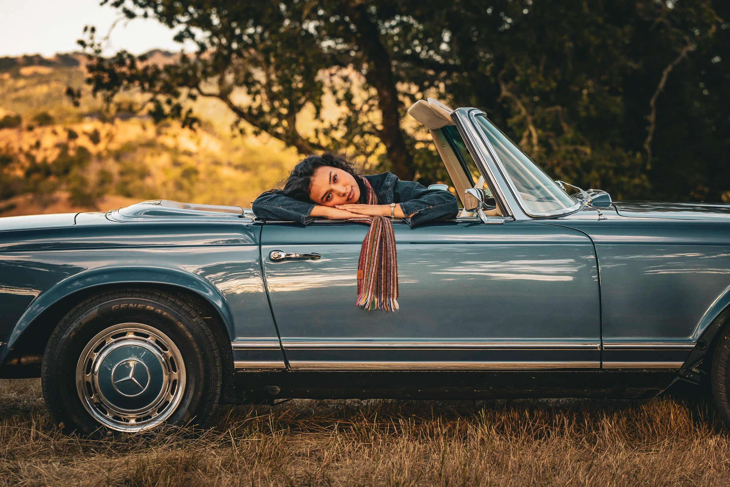 Woman in a denim jacket resting her head on her arms on the door of a vintage blue Mercedes convertible, with a striped scarf hanging down and golden hills behind her