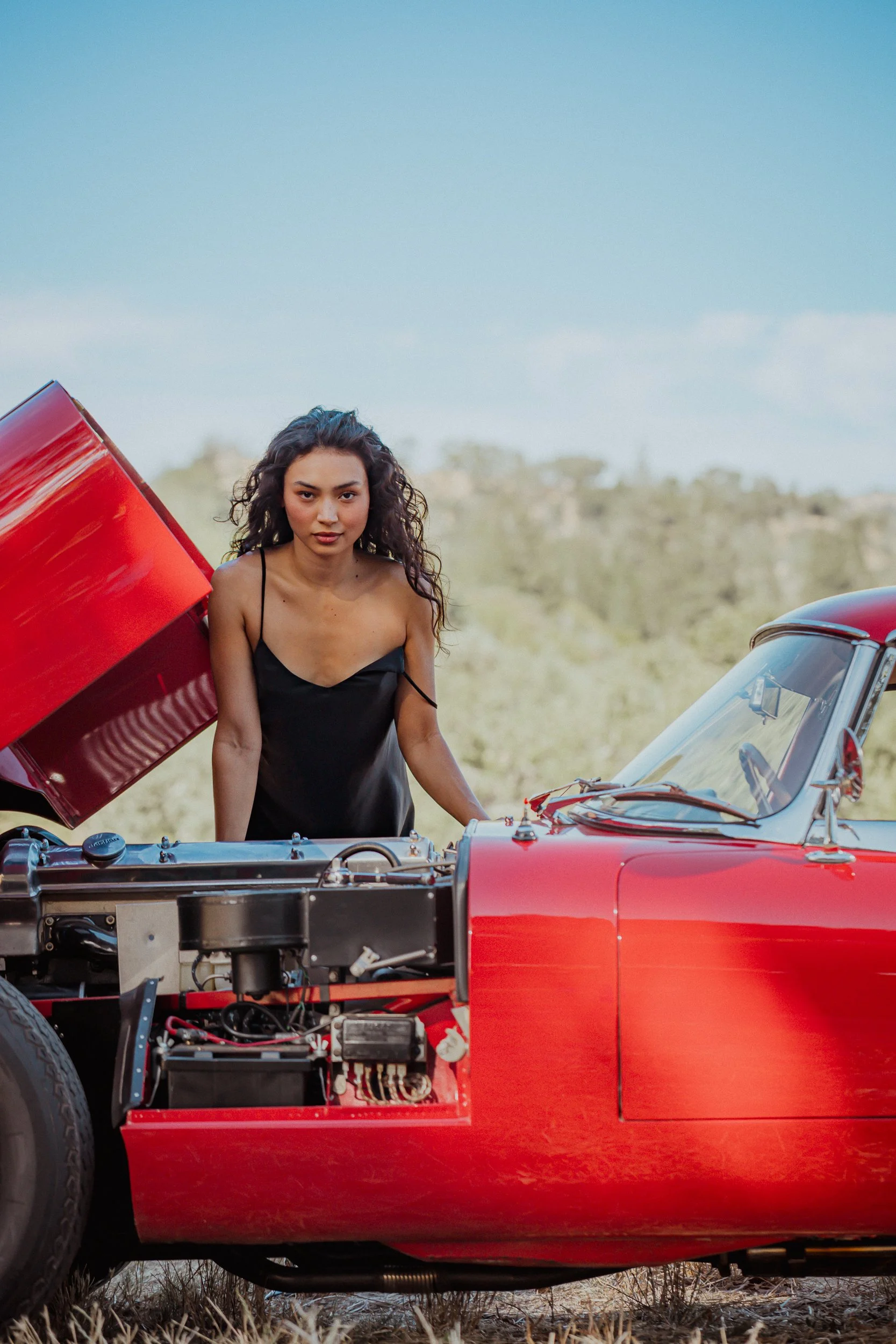 Woman in a black slip dress standing behind the open hood of a vintage red sports car with the engine showing