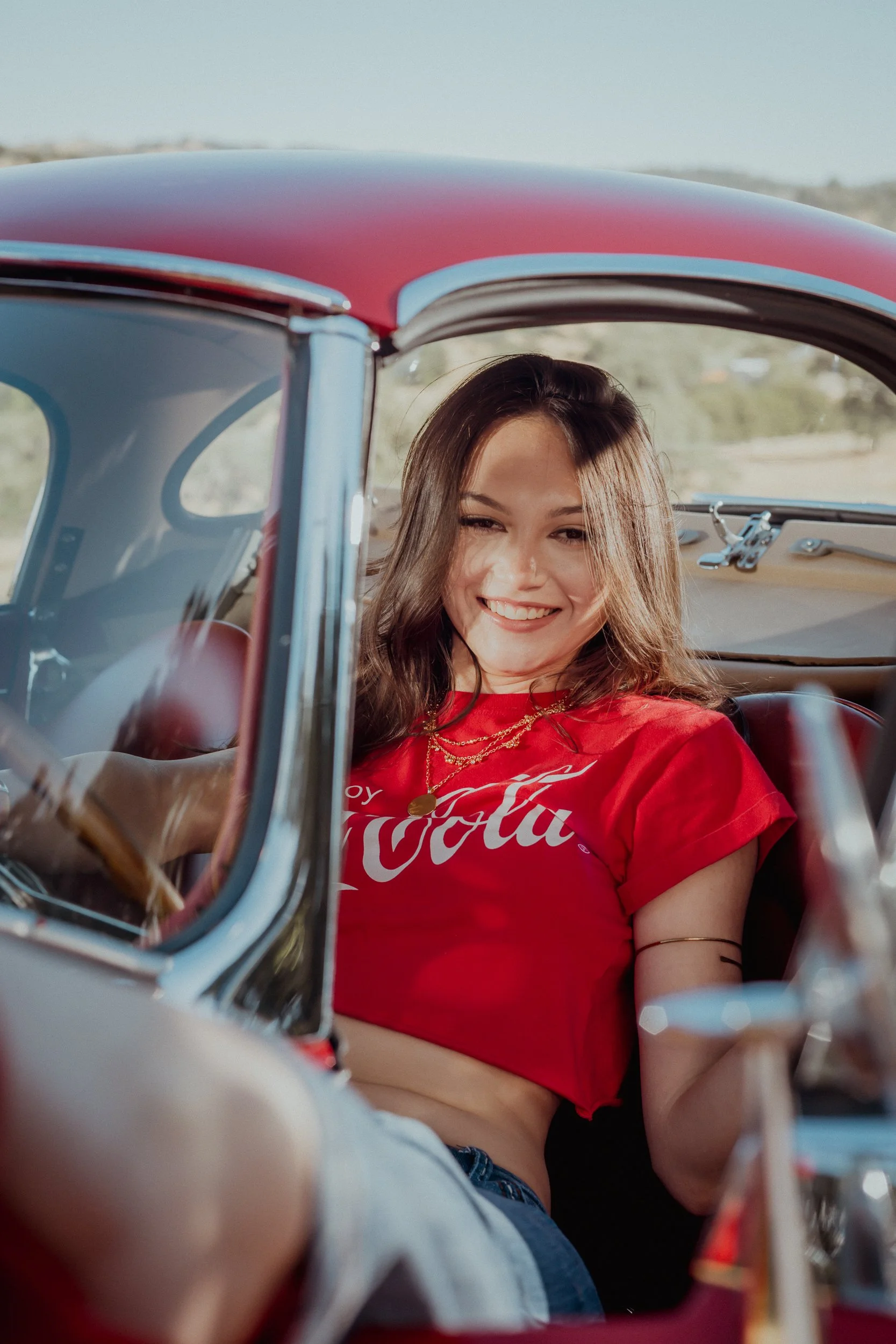 Woman in a red cropped tee looking out while driving a vintage red sports car, seen through the windshield