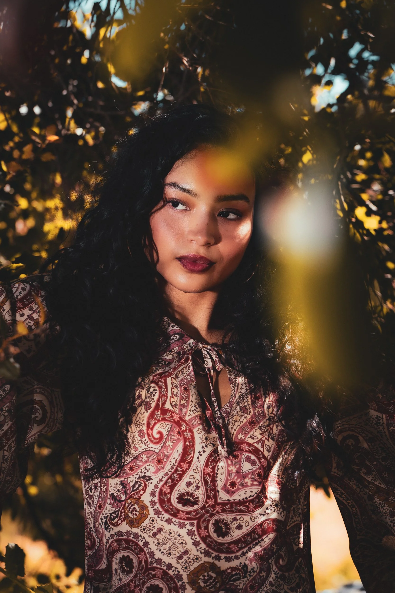 Portrait of a woman with dark curly hair in a red paisley blouse framed by oak branches and golden light