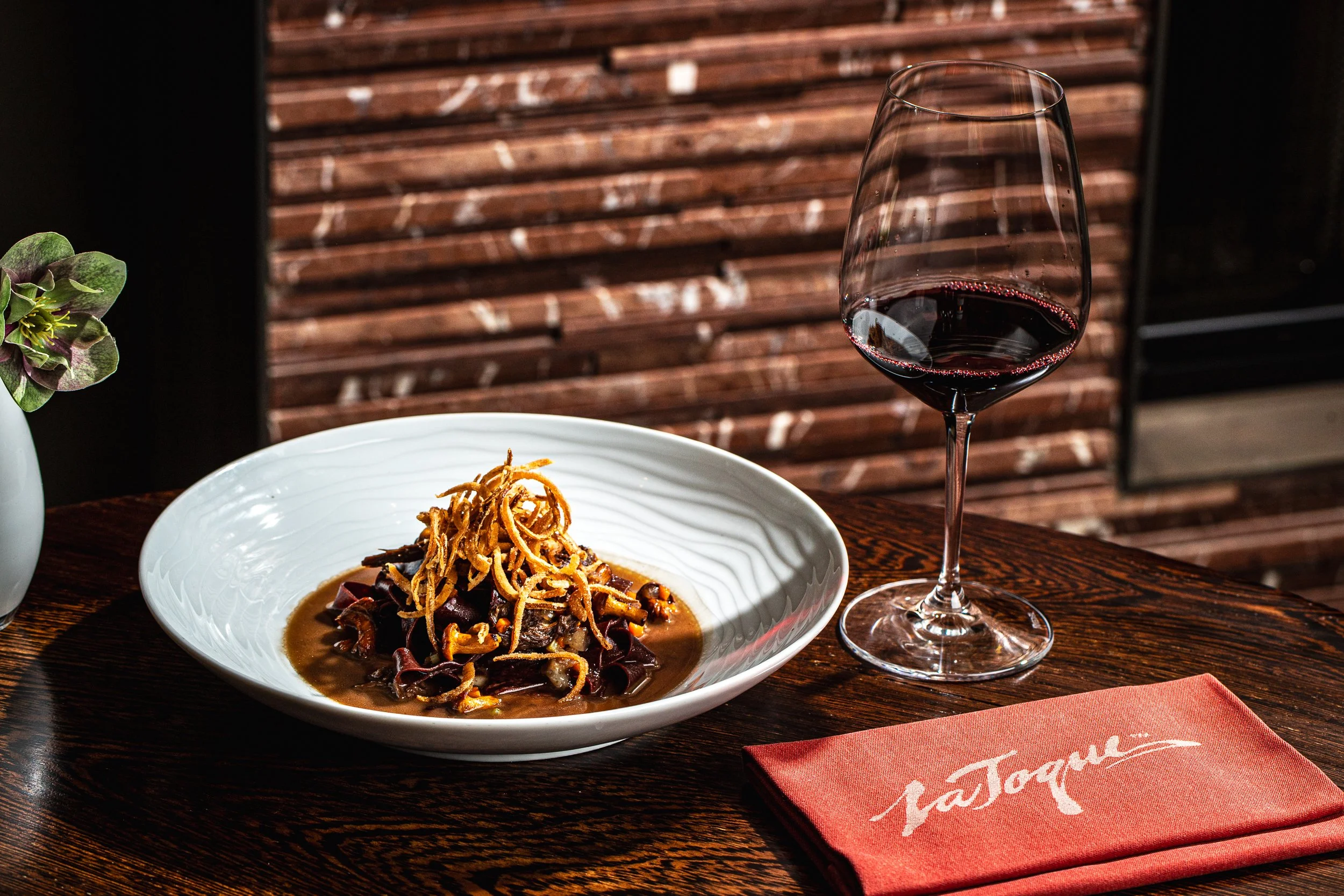 Plated dish topped with crispy shallots beside a glass of red wine and a red La Toque napkin at the restaurant in Napa, California