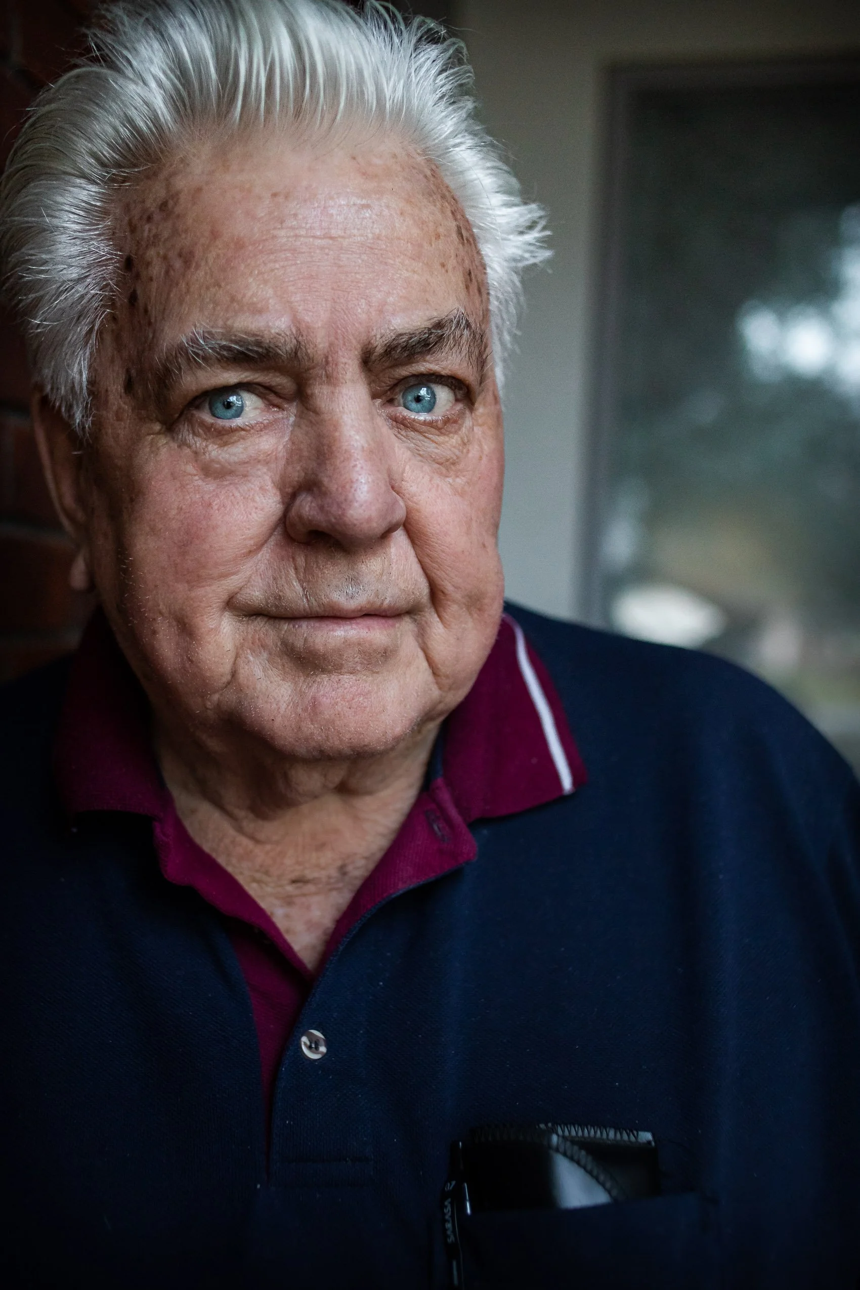Close portrait of an older man with white hair and blue eyes in a navy polo with a burgundy collar
