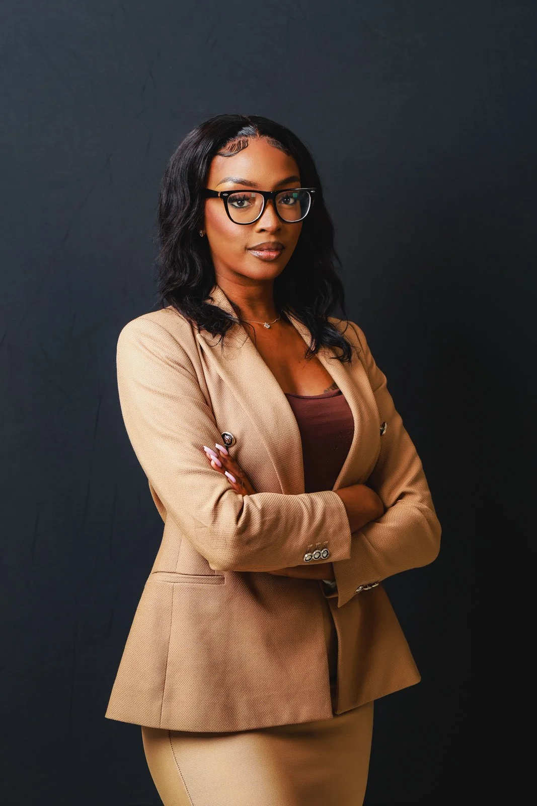 Woman in glasses and a tan blazer standing with her arms crossed against a dark studio backdrop