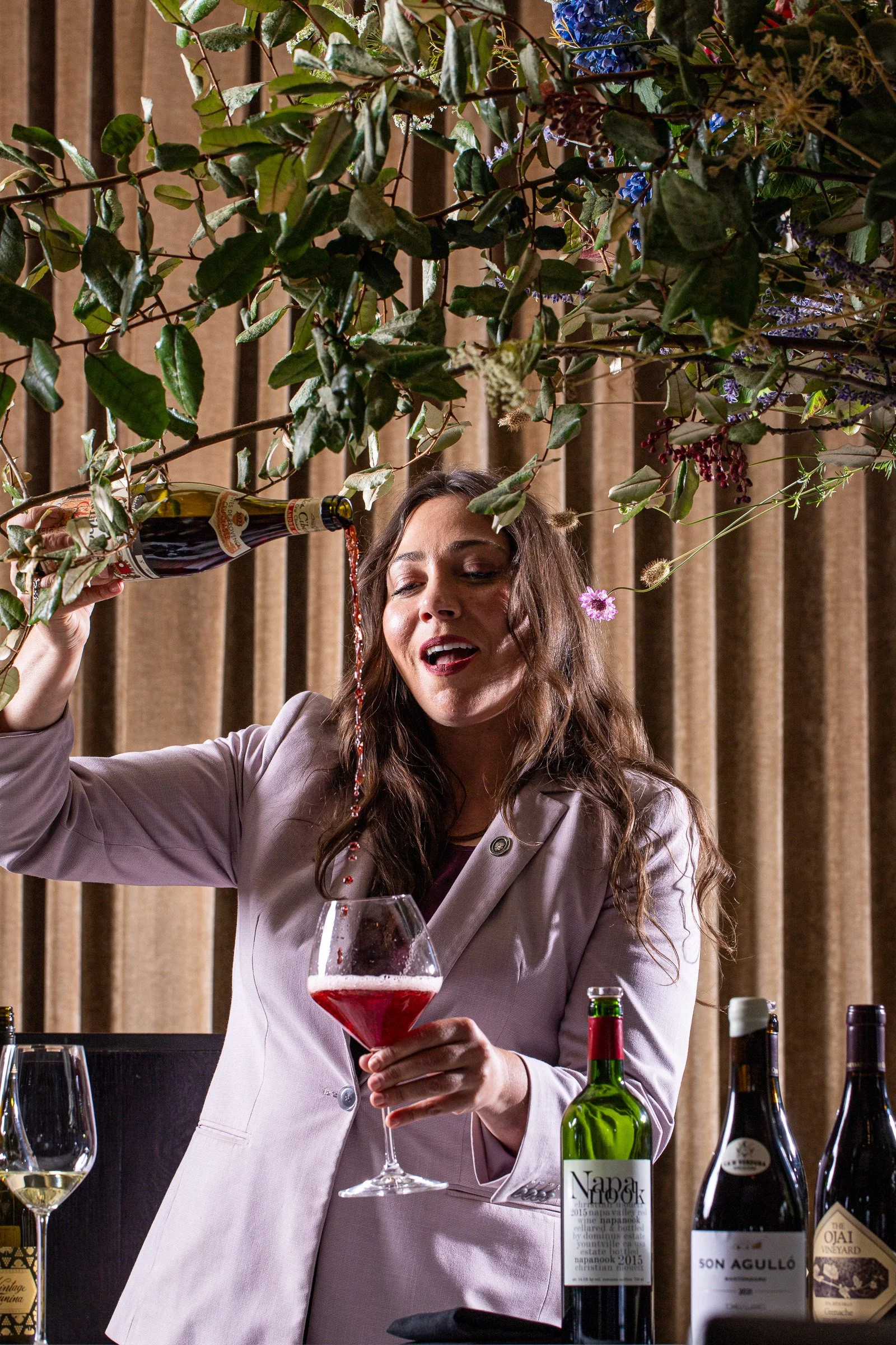 Woman in a lilac blazer laughing as she pours sparkling wine into a glass, with wine bottles in front of her