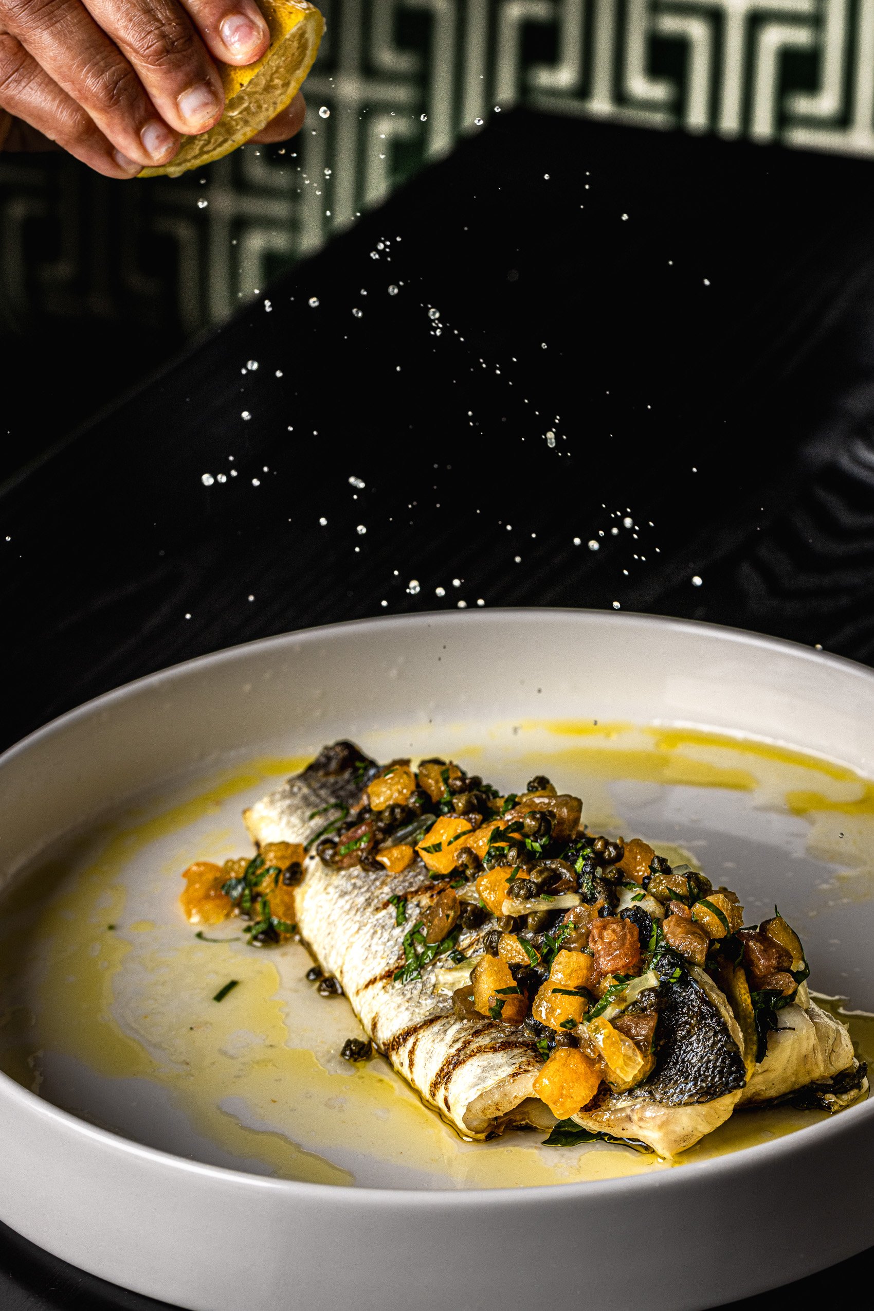 Grilled fish fillet topped with citrus and capers in a white bowl on a black table, with a hand squeezing lemon above it