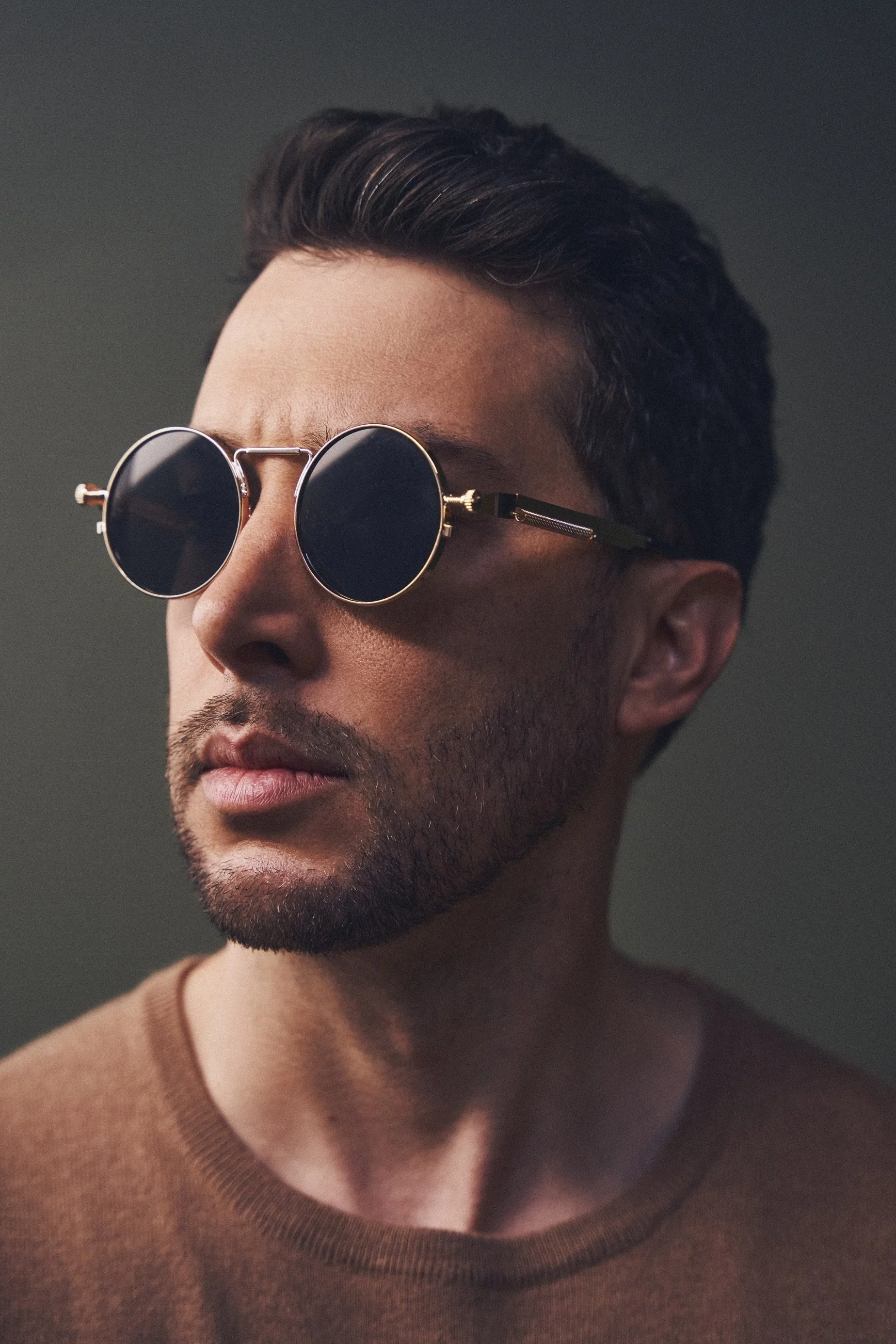 Close-up of a man wearing round black sunglasses, a light brown sweater, with short dark hair and a trimmed beard, against a dark background.