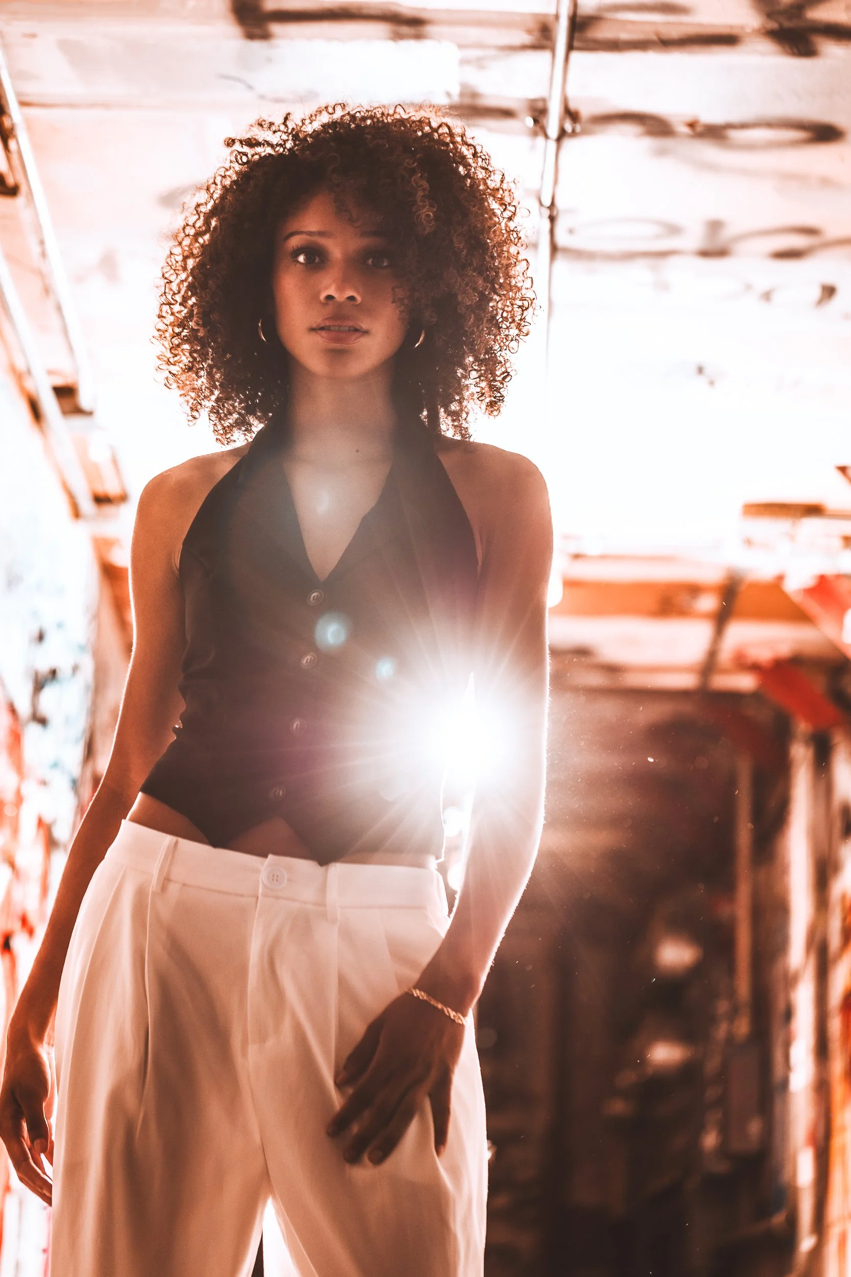 Model in a black halter top and white pants standing in a graffiti tunnel with a bright sun flare behind her
