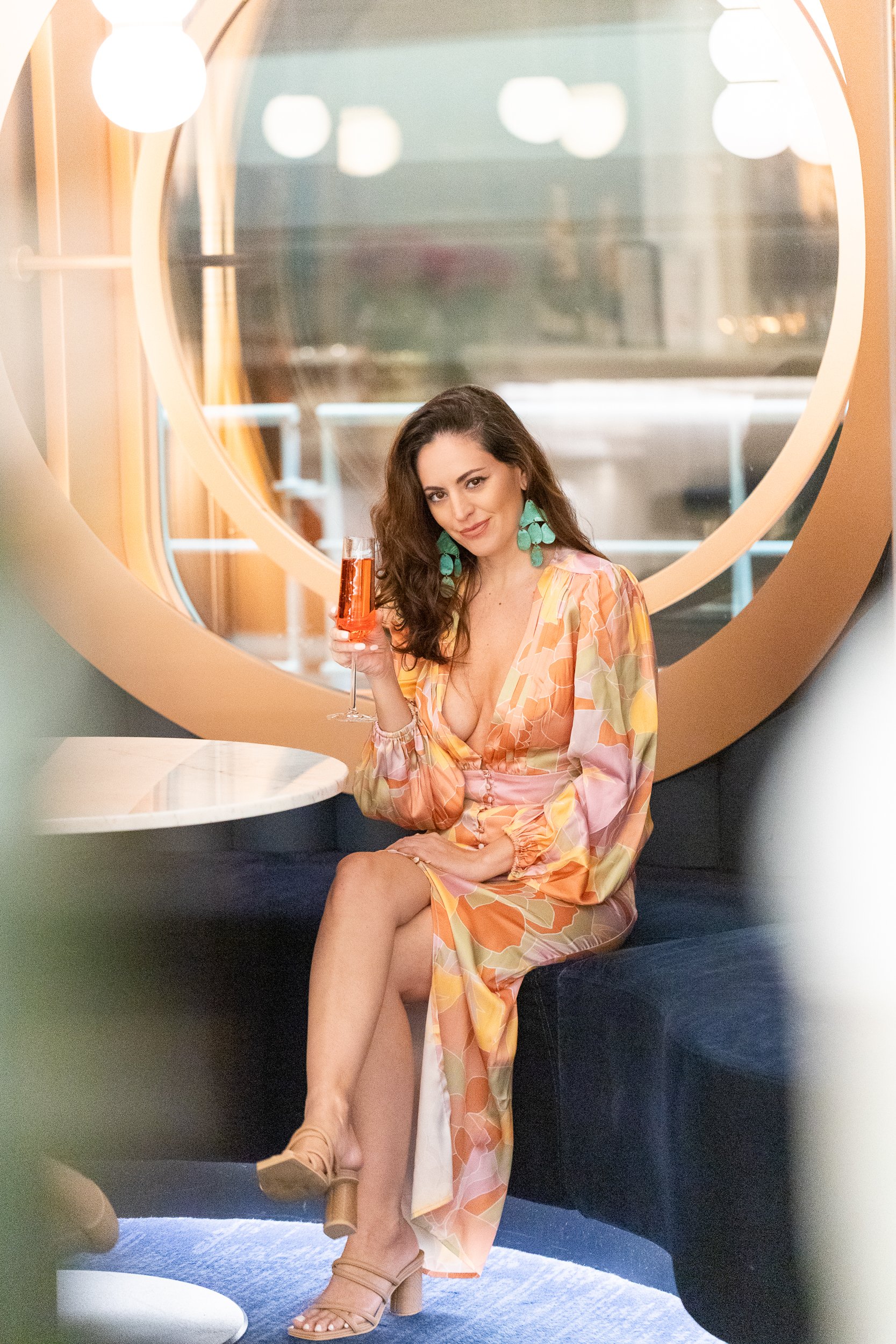Woman in an orange floral robe holding a cocktail, seated in a round mirrored nook