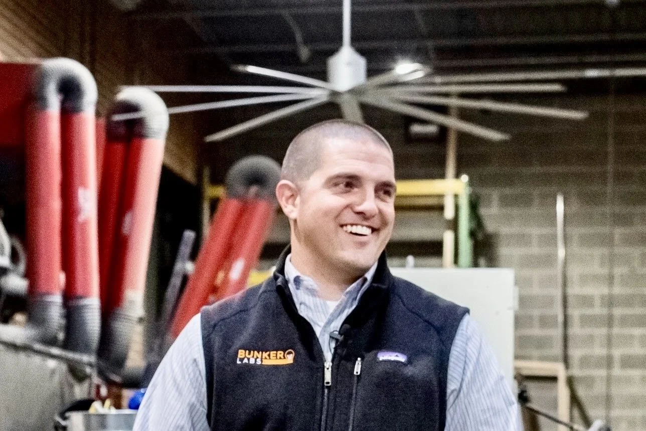 A man smiling in an industrial setting with tools and equipment, including red pipes and a ceiling fan.