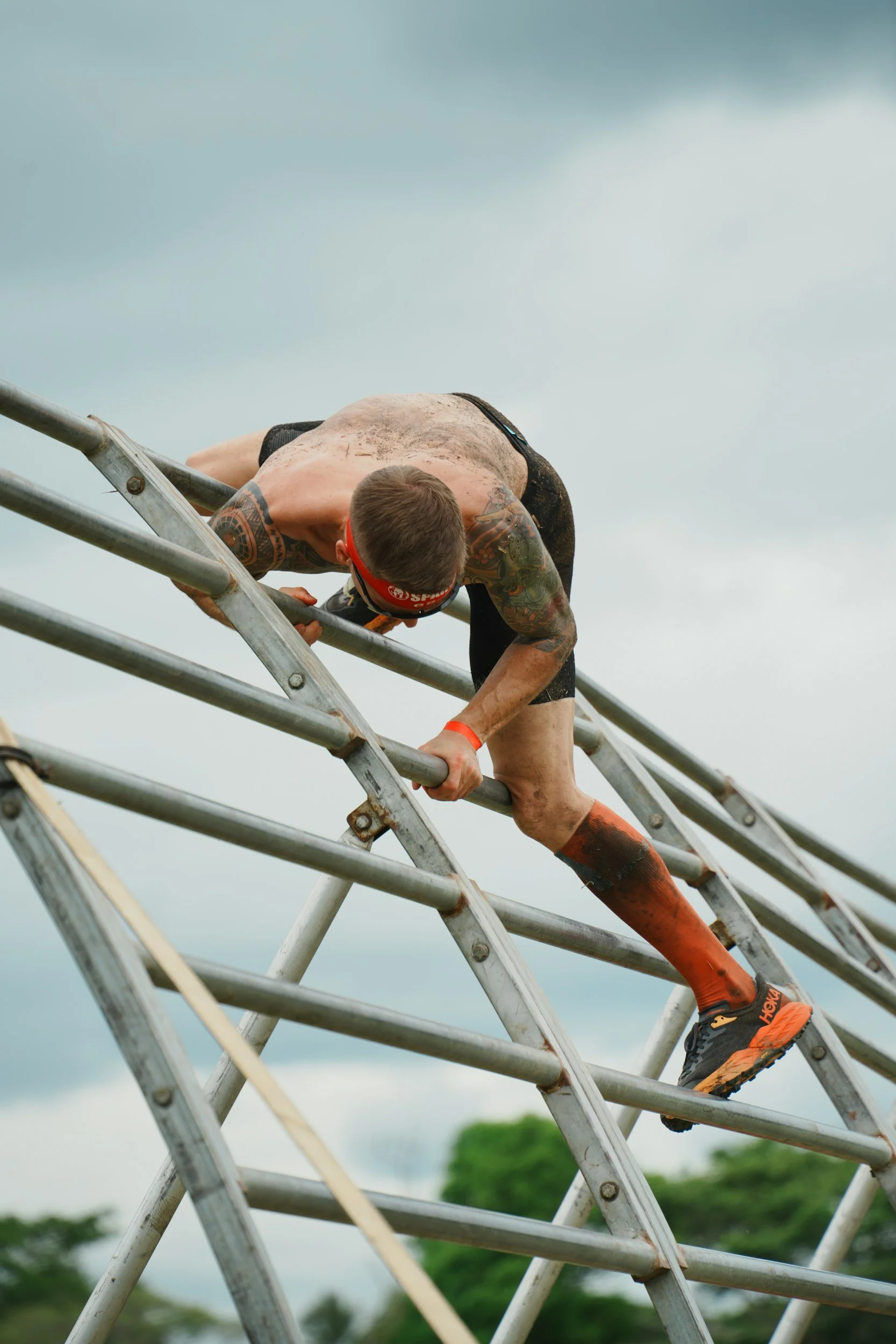 A person climbing a metal obstacle during an outdoor race, wearing athletic gear and a red headband, with cloudy sky and trees in the background.