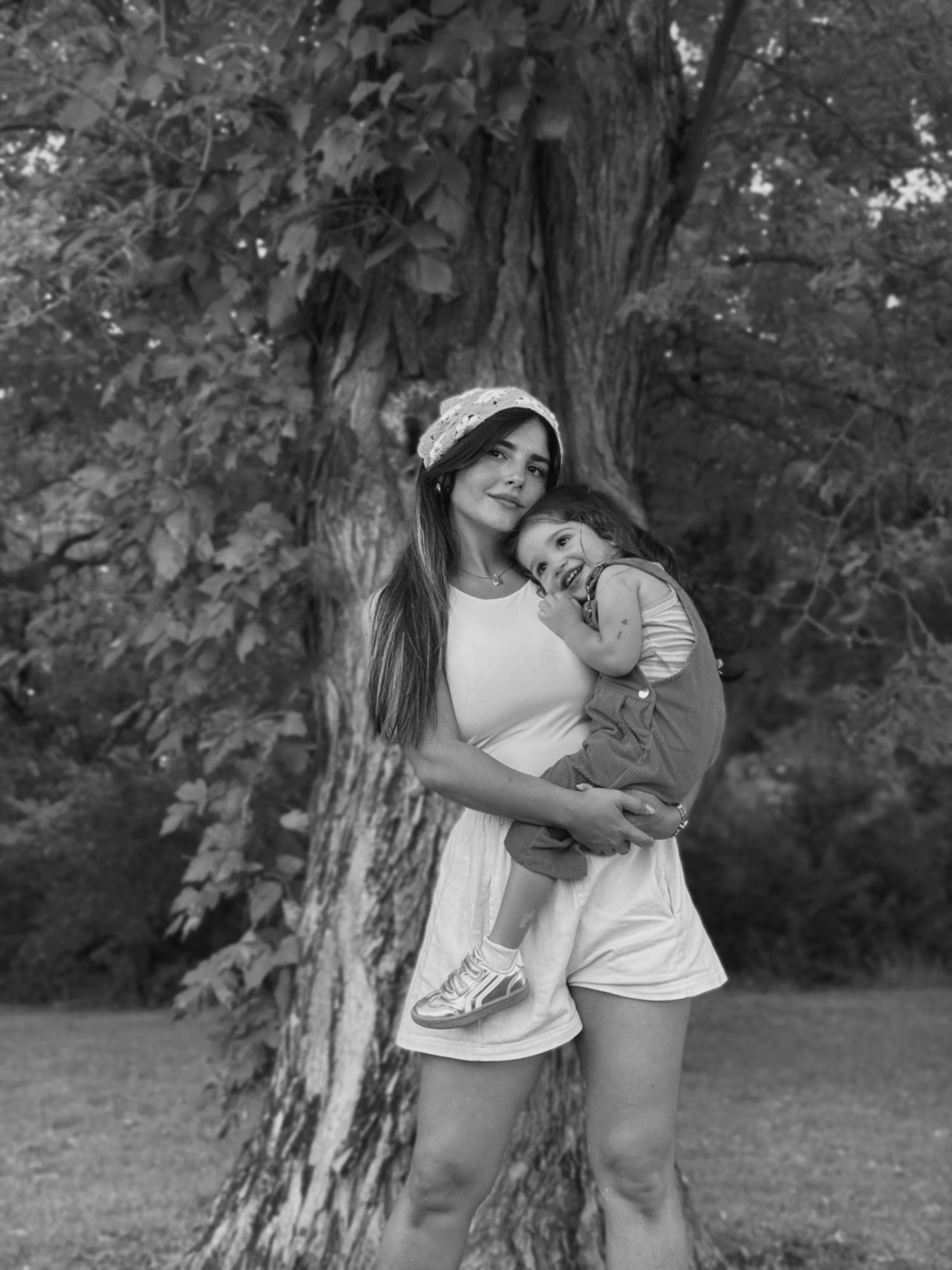 A woman holding a young girl outdoors in front of a large tree with broad leaves, in black and white photograph.