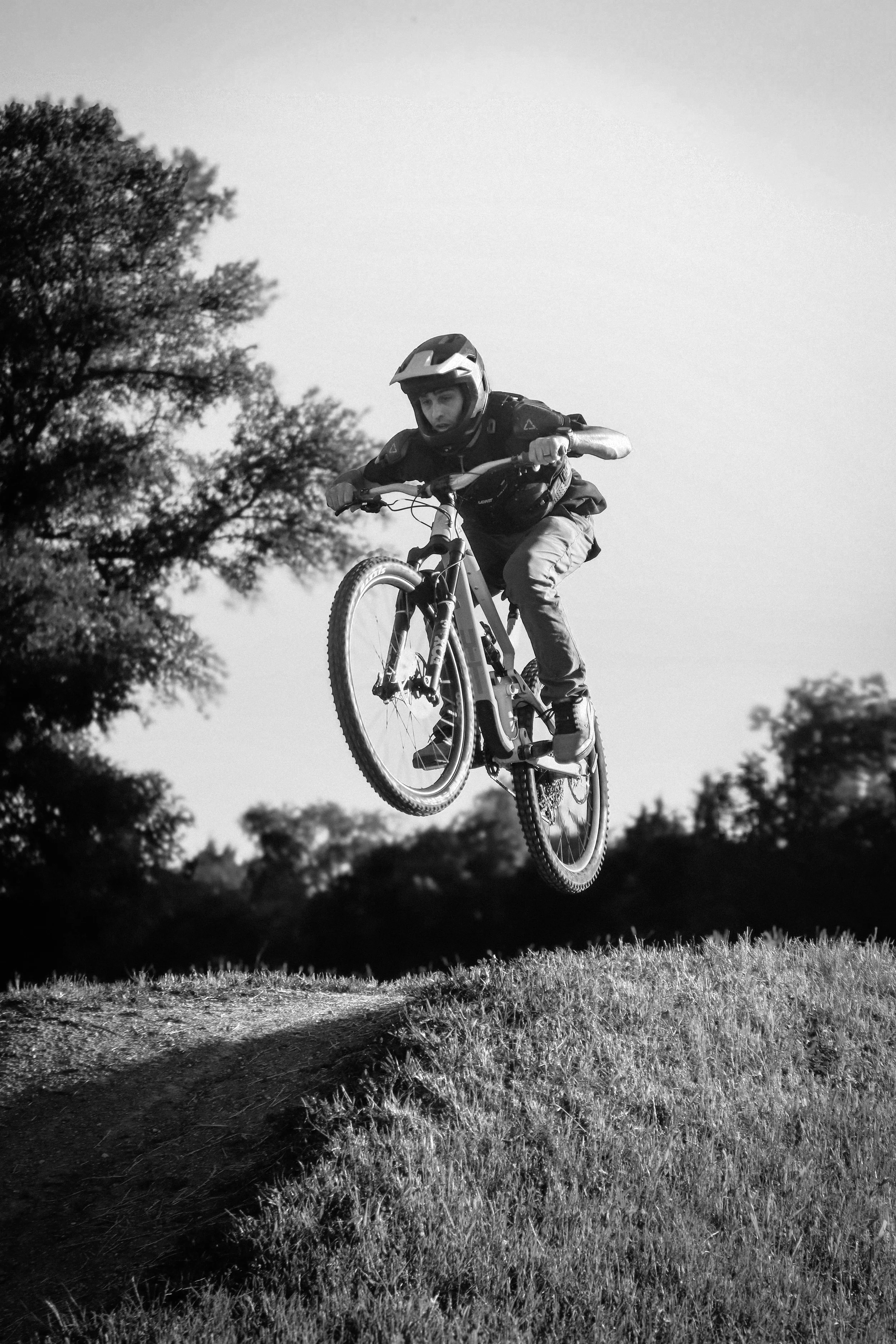 A person riding a mountain bike and jumping in mid-air in a grassy outdoor area, wearing a helmet and protective gear.
