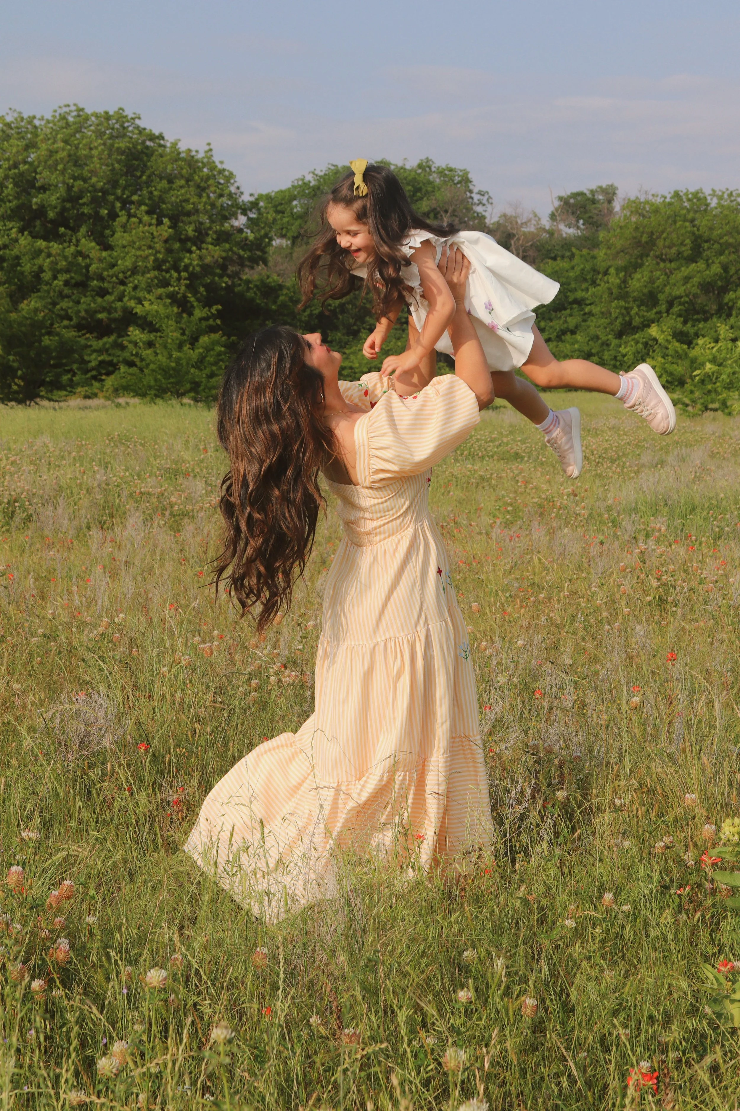 A woman lifts a young girl into the air in a grassy field with green trees in the background, both smiling and enjoying a joyful moment.