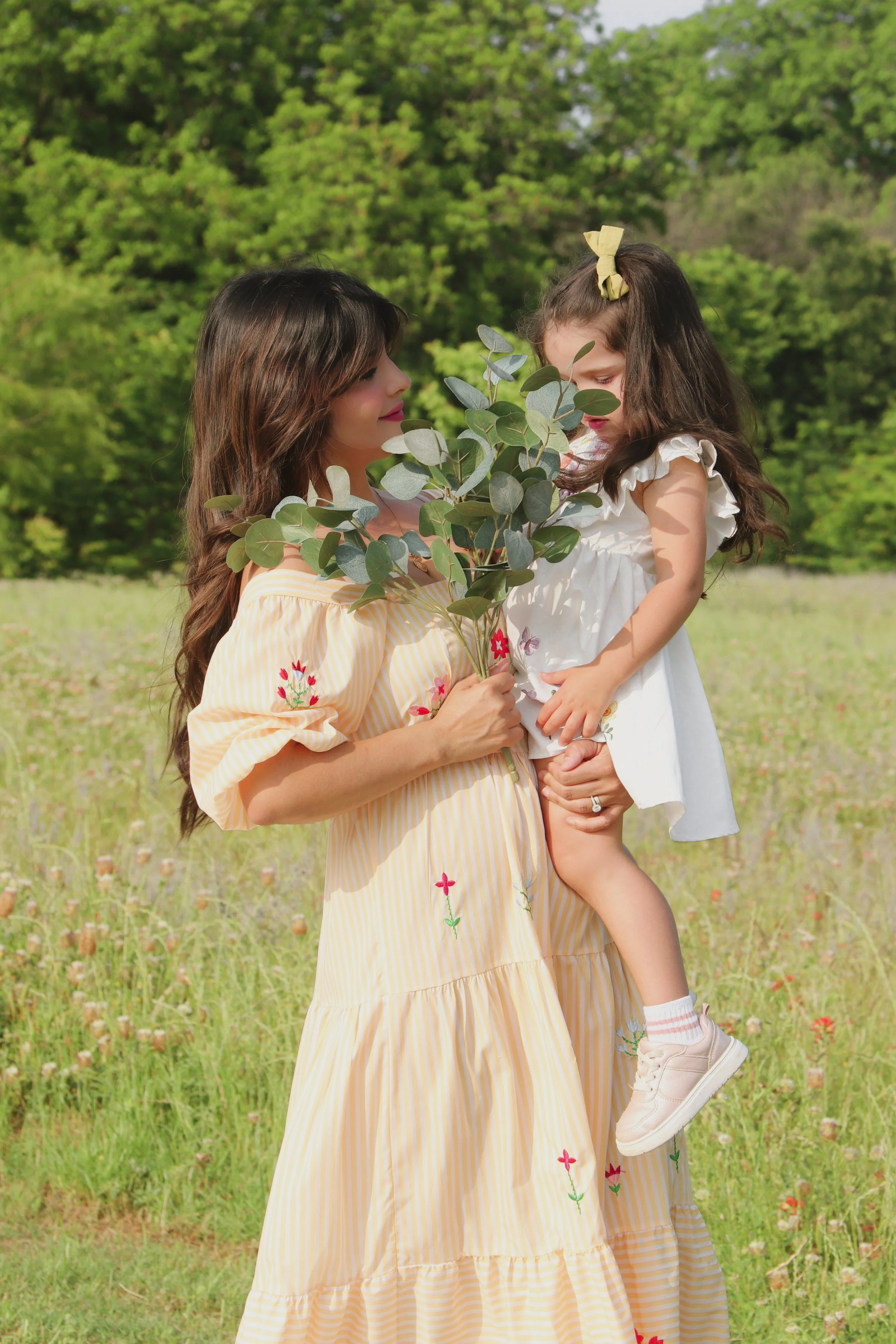 A woman holding a young girl in a grassy field surrounded by trees, with the woman carrying a bouquet of green leaves.
