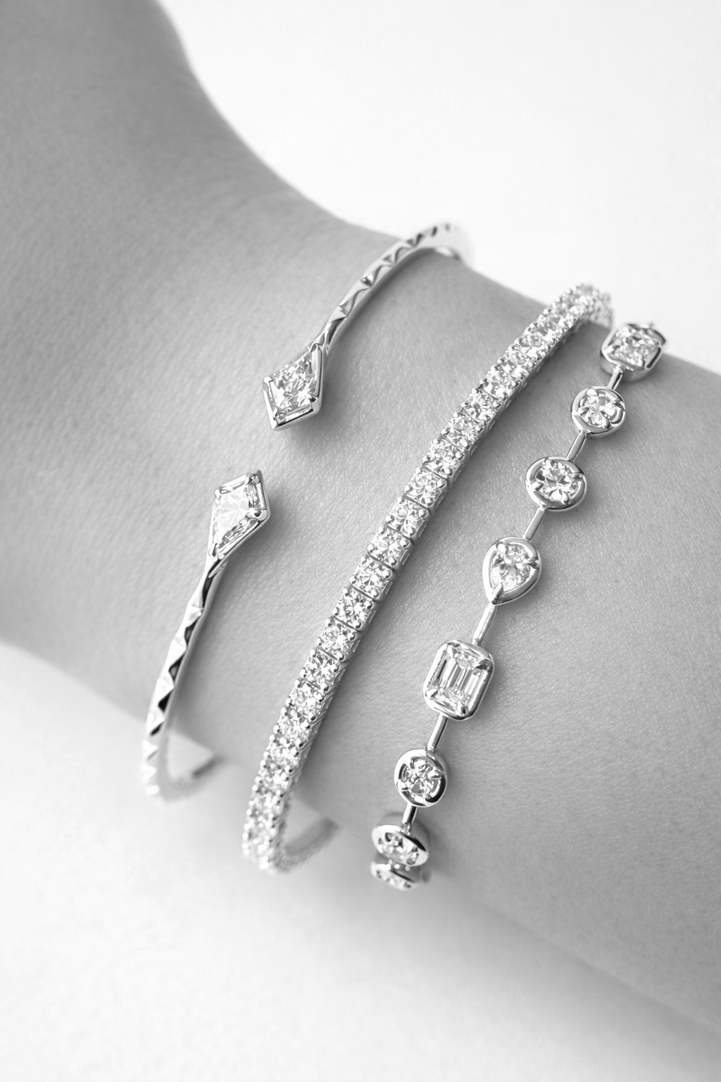 Three silver and diamond bracelets displayed on a person's wrist, featuring geometric and round-cut stones.