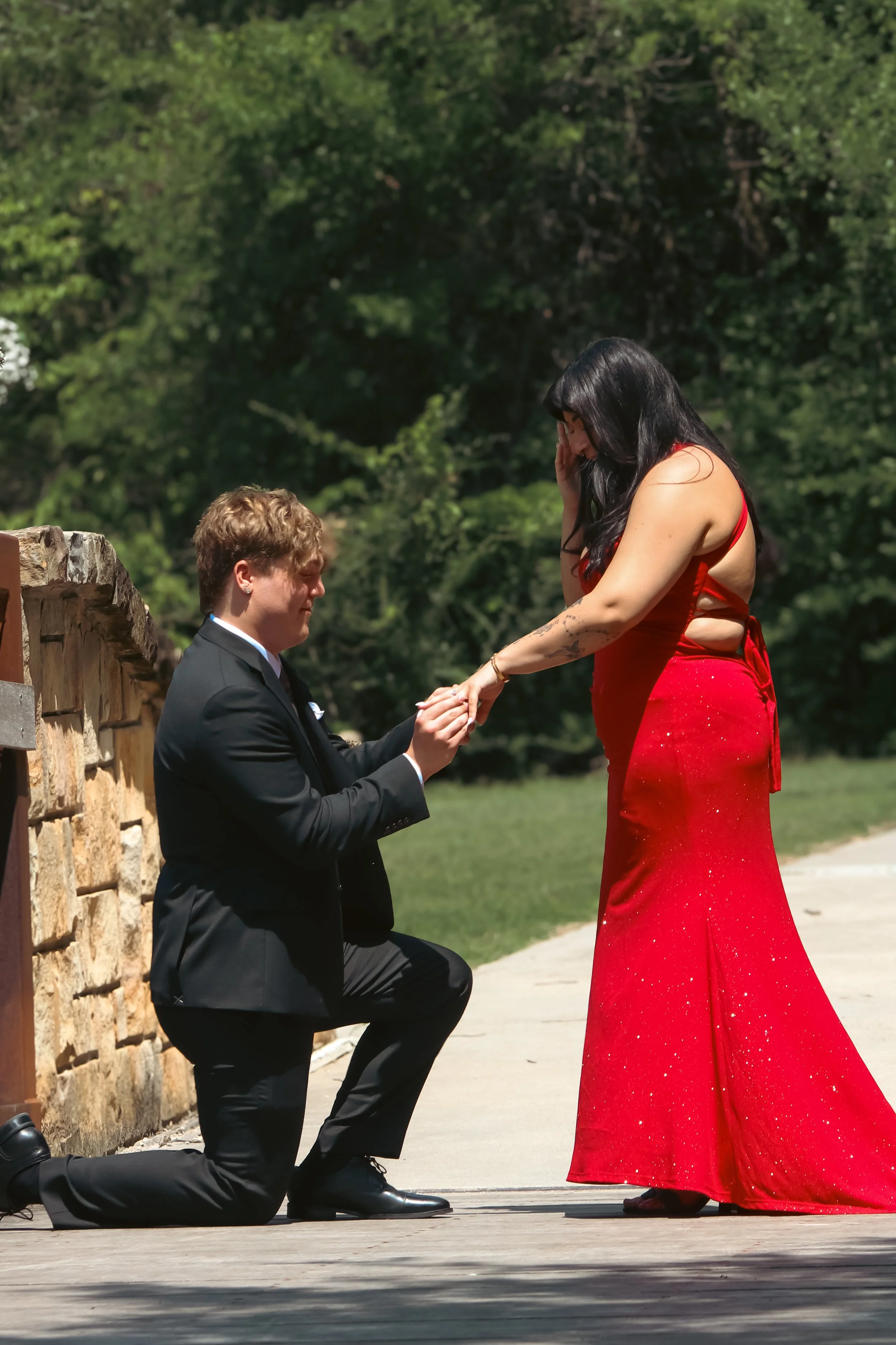 A man dressed in a black suit is kneeling on one knee and proposing marriage to a woman in a long red dress. The woman appears emotional, covering part of her face with her hand, while the man holds her hand.